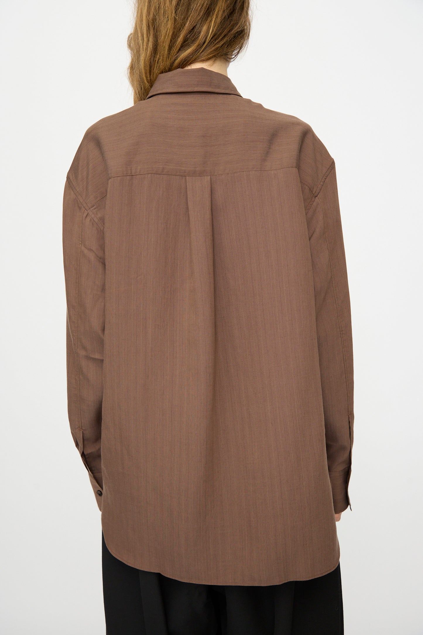 A person stands with their back to the camera, wearing the Studio Nicholson Subtle Pinstripe Leto Shirt in brown and black pants against a plain white background.