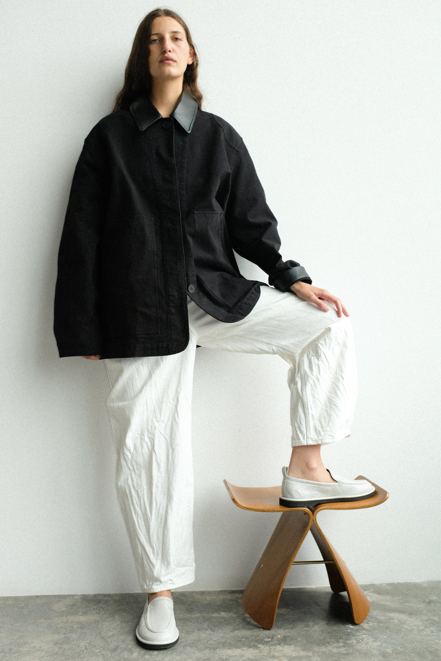 Person wearing a black chore jacket and white pants with one foot on a wooden stool against a white wall.