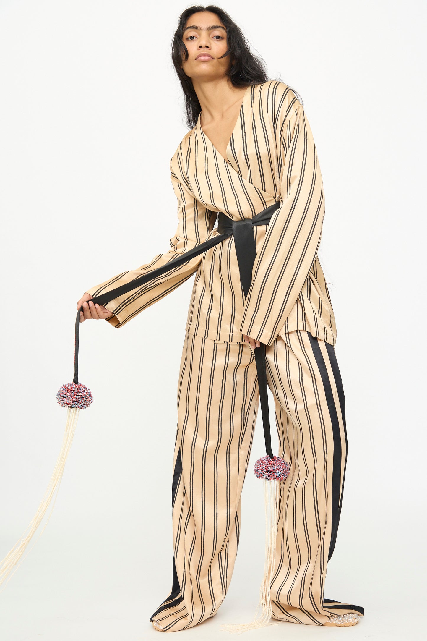 A woman stands against a plain white background, wearing wide-leg beige pants with black vertical stripes and accessorizing her outfit with the TIGRA TIGRA Pillows Belt featuring a glass bead fringe.