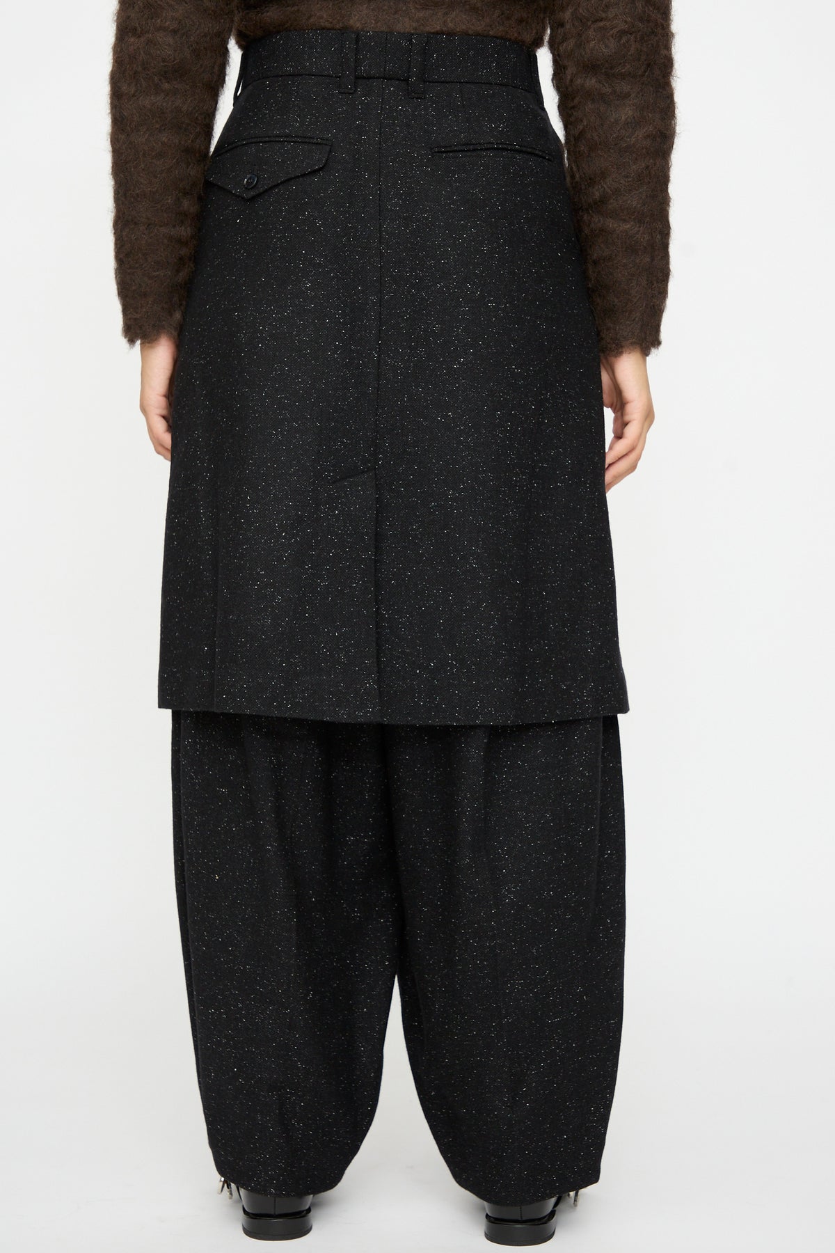 A person is seen from the back, wearing a black speckled wool skirt over TOGA ARCHIVES Layered Trousers in Black, paired with a brown textured long-sleeve top.