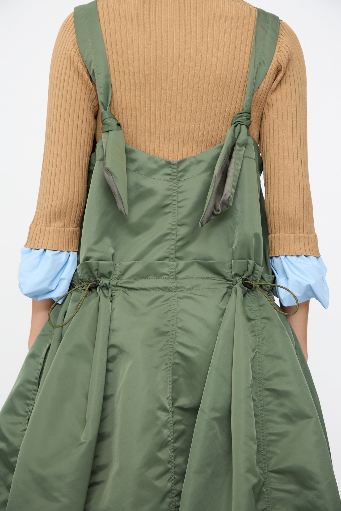 A person wearing a tan ribbed sweater with light blue cuffs, layered under the TOGA ARCHIVES Nylon Twill Dress in Khaki—an A-line, cinched, sleeveless style—viewed from the back.