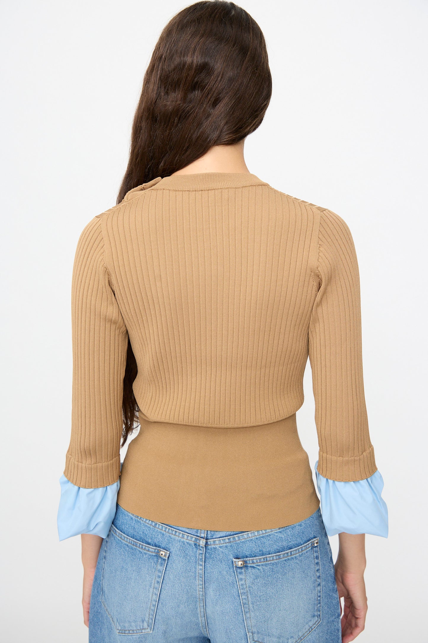 A woman with long brown hair is seen from the back wearing the TOGA ARCHIVES Rib Knit Top in Camel, which features a slim fit and blue cuffs, paired with light blue jeans.
