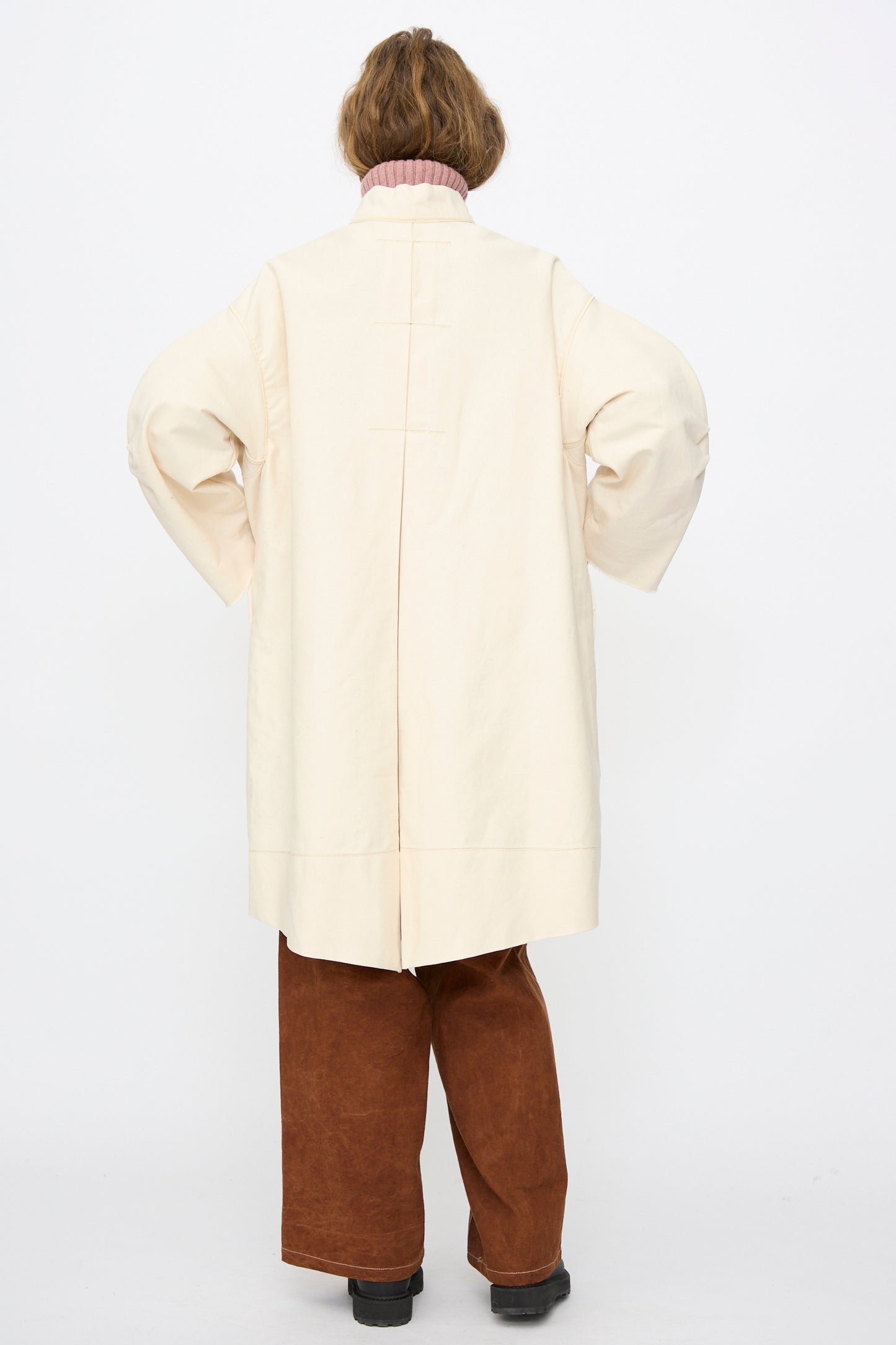 Person standing with their back to the camera, wearing the Thank You Have A Good Day Denim Fishtail Parka in Ecru, brown pants, black shoes, and a pink turtleneck.