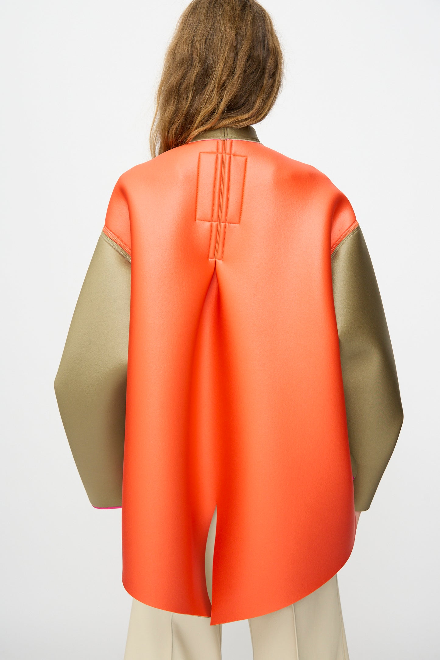 A person faces away from the camera, wearing the Thank You Have A Good Day Color block Cropped Parka with an orange cape-like panel over beige, paired with off-white pants.