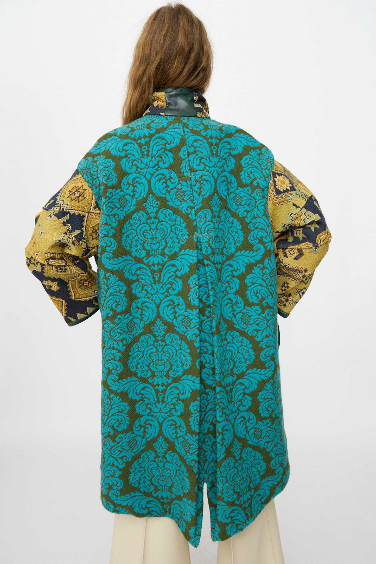 A person with long hair is seen from the back, wearing the Thank You Have A Good Day Patchwork Fishtail Parka, featuring a teal and green pattern with gold and black tapestry-inspired sleeves, paired with light-colored pants.