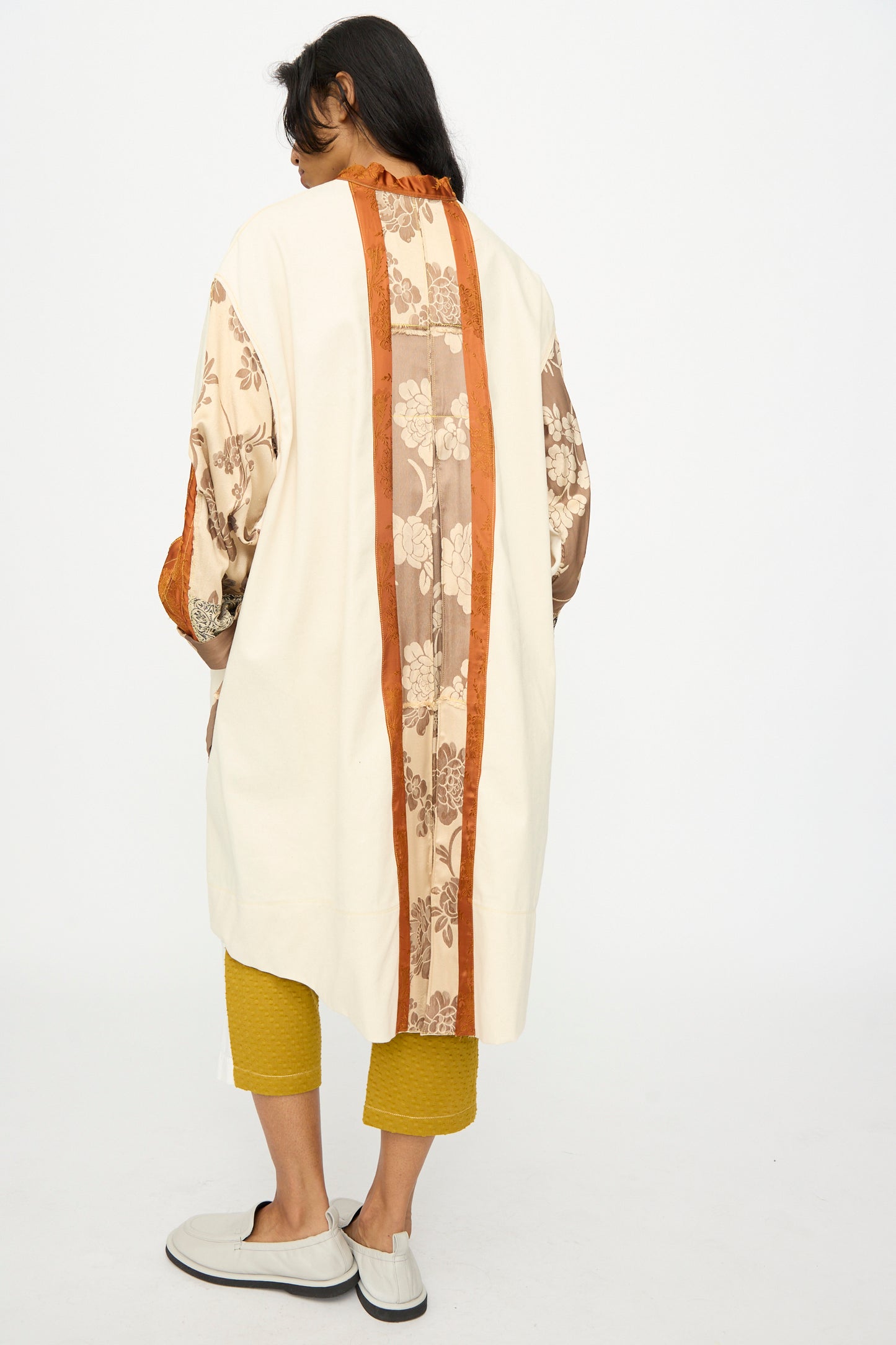 A person faces away in Thank You Have A Good Day’s Patchwork Parka—cream with brown and orange floral panels—paired with yellow cropped pants and light slip-ons, featuring vintage buttons and handmade in Tribeca.