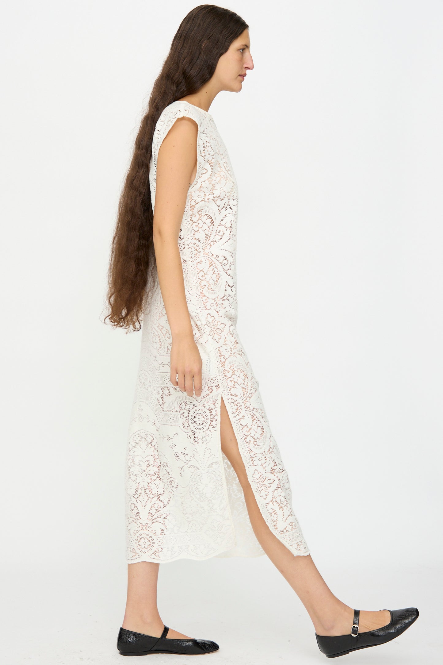 A person with long brown hair models the Quaker Lace Caftan by Thank You Have A Good Day, a sleeveless white lace dress with a side slit, paired with black flats, shown in profile against a plain white background.