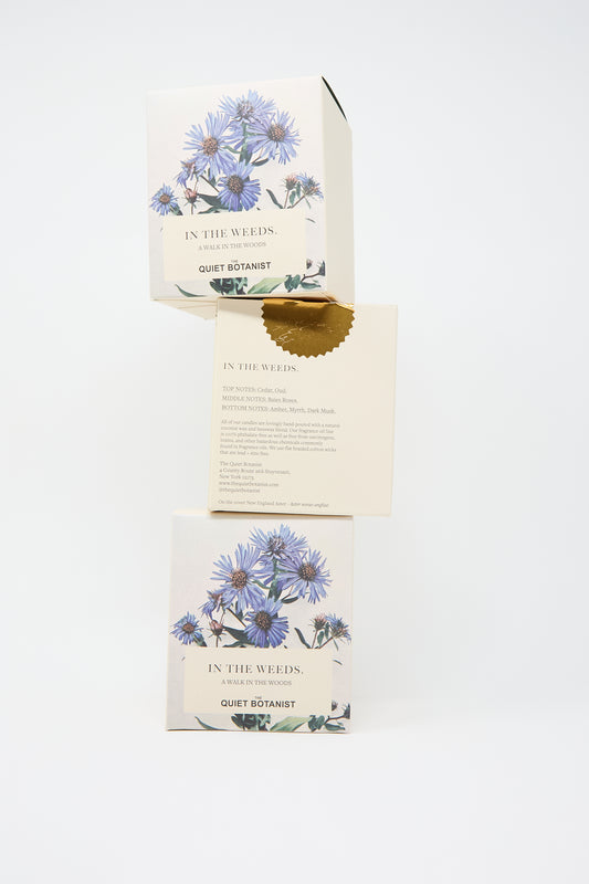 Three stacked boxes of In the Weeds Candle by The Quiet Botanist. Made with natural coconut wax, the packaging features botanical illustrations and a gold seal.