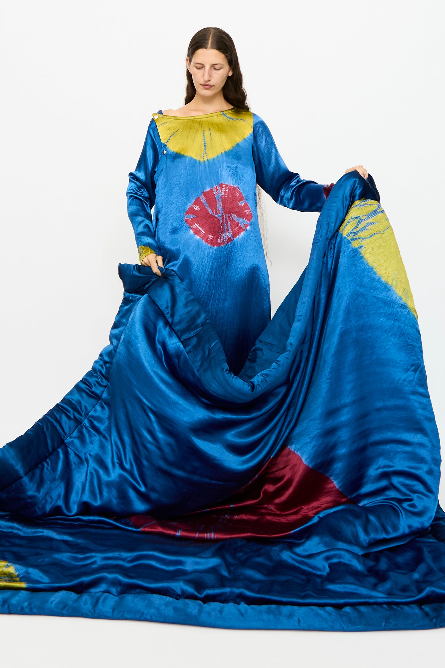 A person in a long, jewel blue gown with acid green and magenta patterns stands against a white background, holding the dress inspired by TIGRA TIGRA’s Moon Dye Quilt.