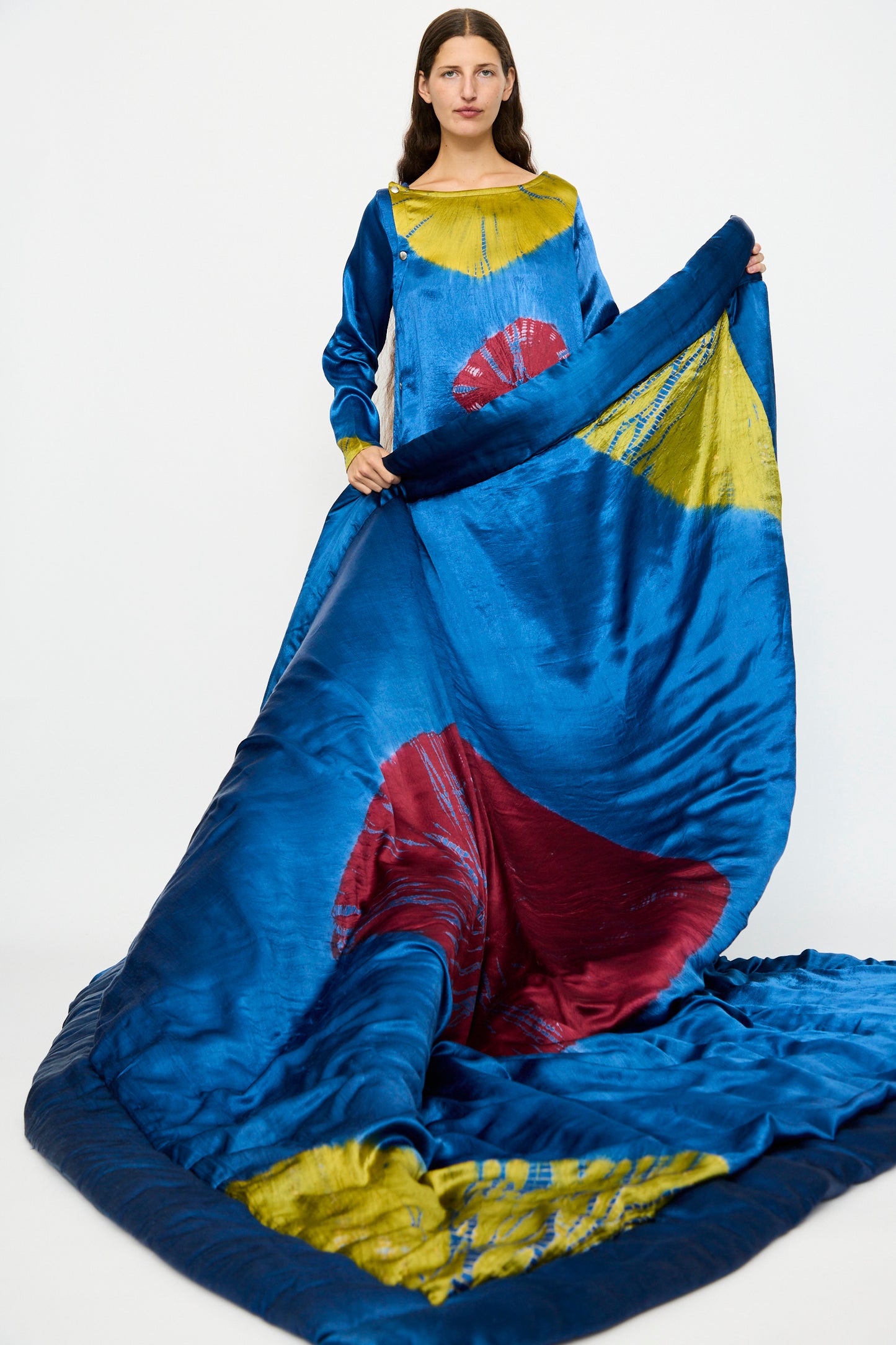 A woman stands in front of a plain white background wearing the TIGRA TIGRA Moon Dye Quilt in Jewel Blue, Acid Green, and Magenta, with its long, flowing fabric and bold circular patterns extended outward.