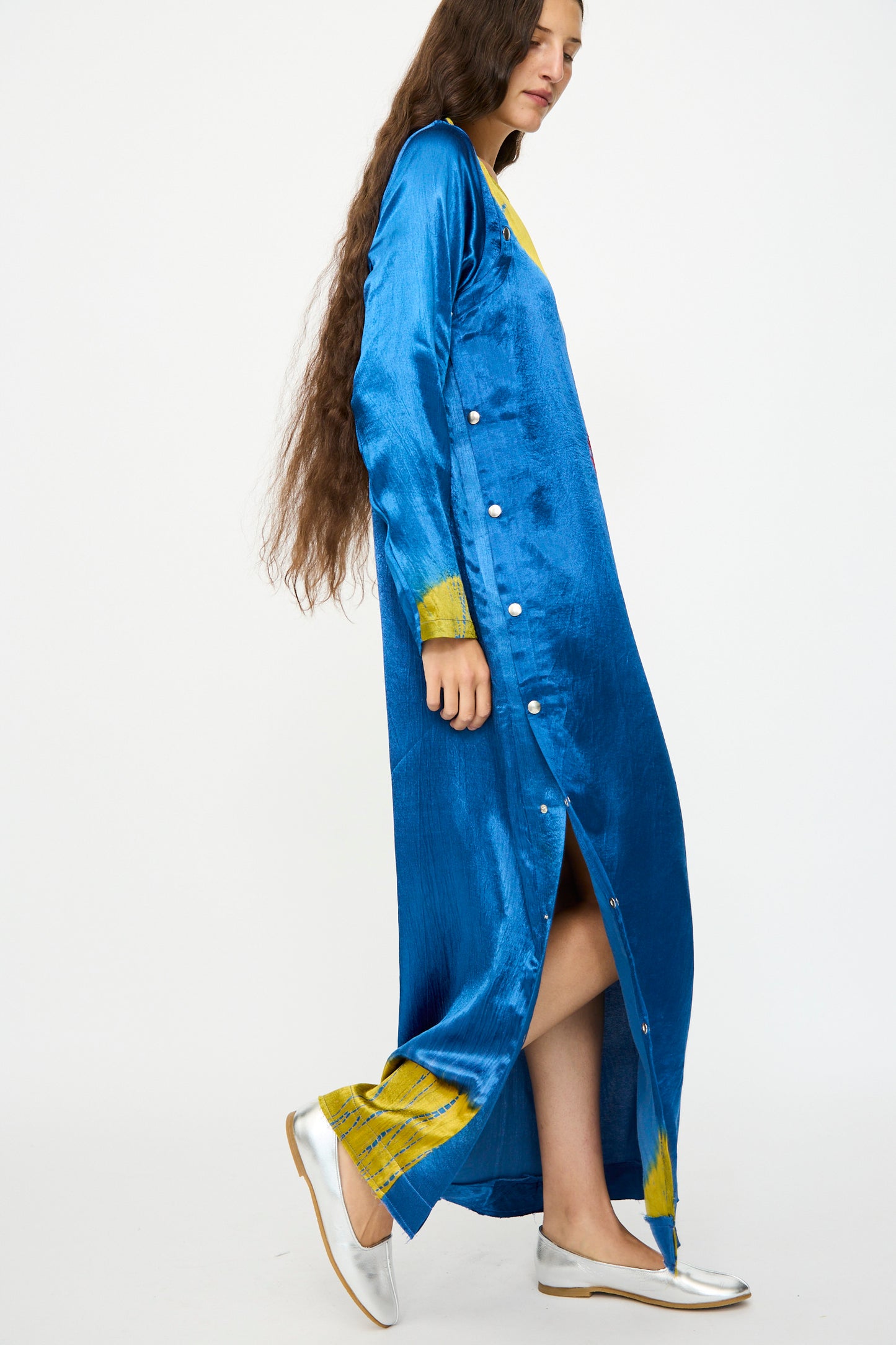 A person with long brown hair stands against a plain white background wearing the TIGRA TIGRA Shibori Snap Dress in Jewel Blue, Acid Green, and Magenta, styled with white flats.