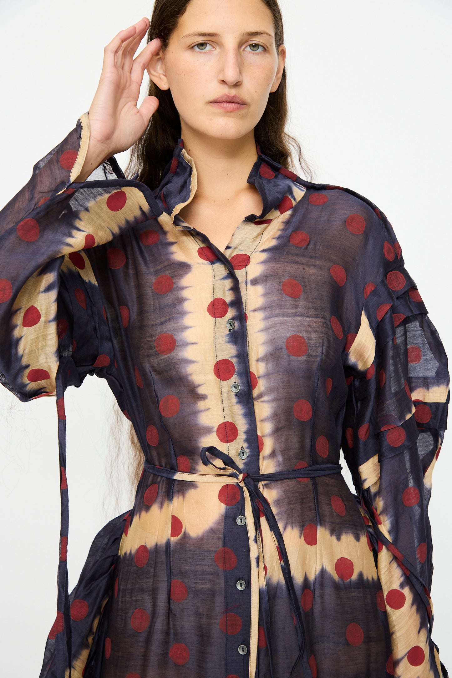 A woman wearing the TIGRA TIGRA Silk Chanderi Teatime Dress in navy, cream, and red—featuring sheer fabric with polka dots, subtle tie-dye patterns, front buttons, and a slim waist tie—stands against a plain background.