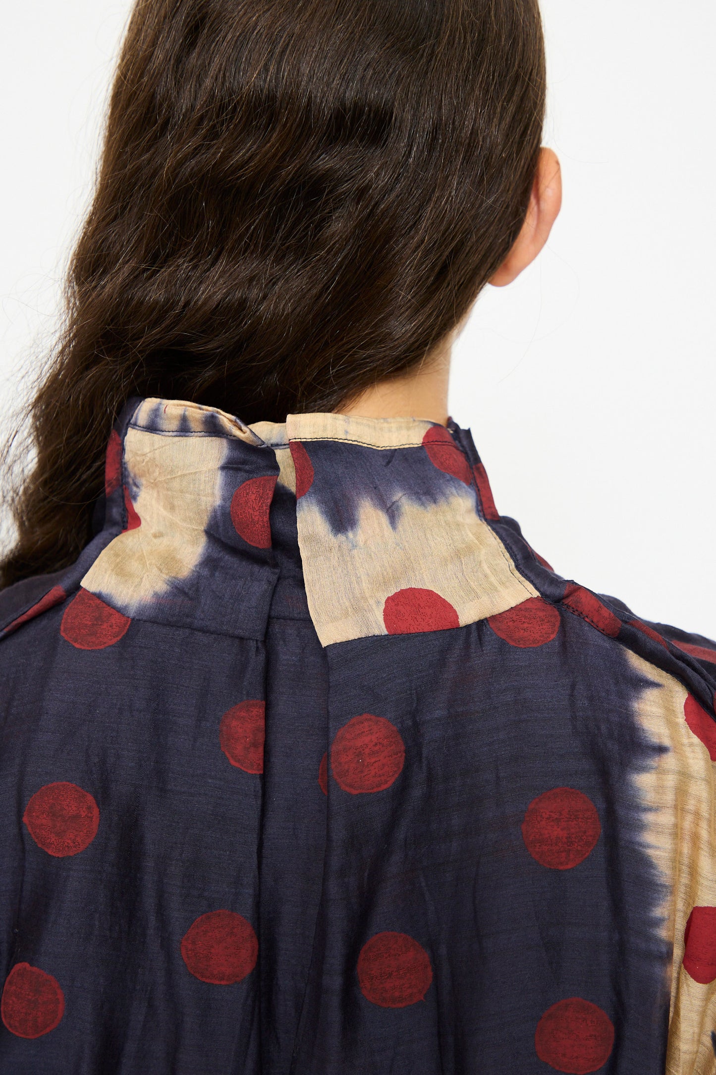 Back view of a person with long dark hair wearing the TIGRA TIGRA Silk Chanderi Teatime Dress in navy, cream, and red, featuring polka dots and a high collar with beige and dark blue tie-dye pattern.
