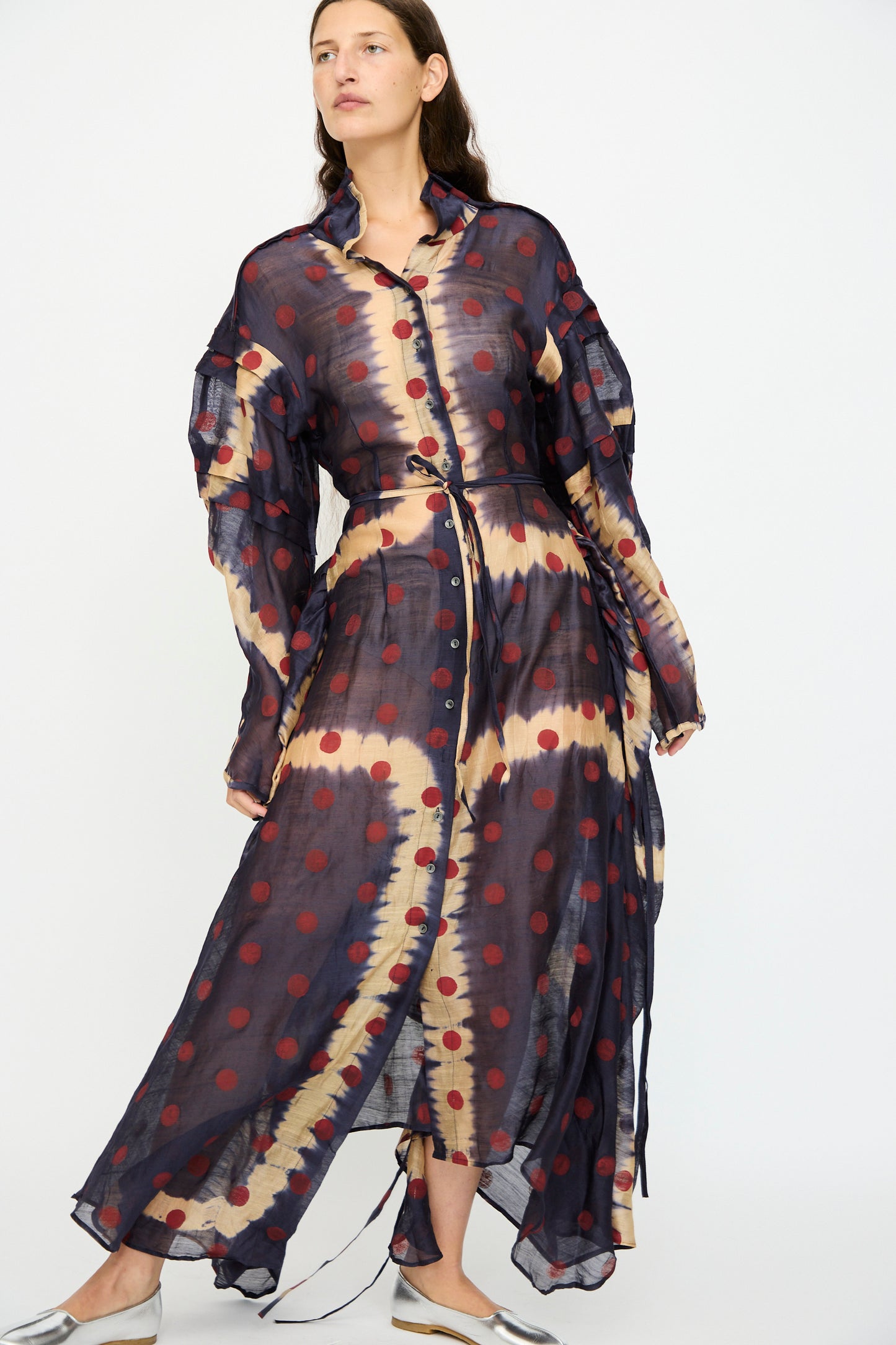 A woman wears the TIGRA TIGRA Silk Chanderi Teatime Dress in Navy, Cream and Red, featuring a sheer maxi silhouette with red polka dots, a collar, and front buttons, paired with metallic flats against a plain background.