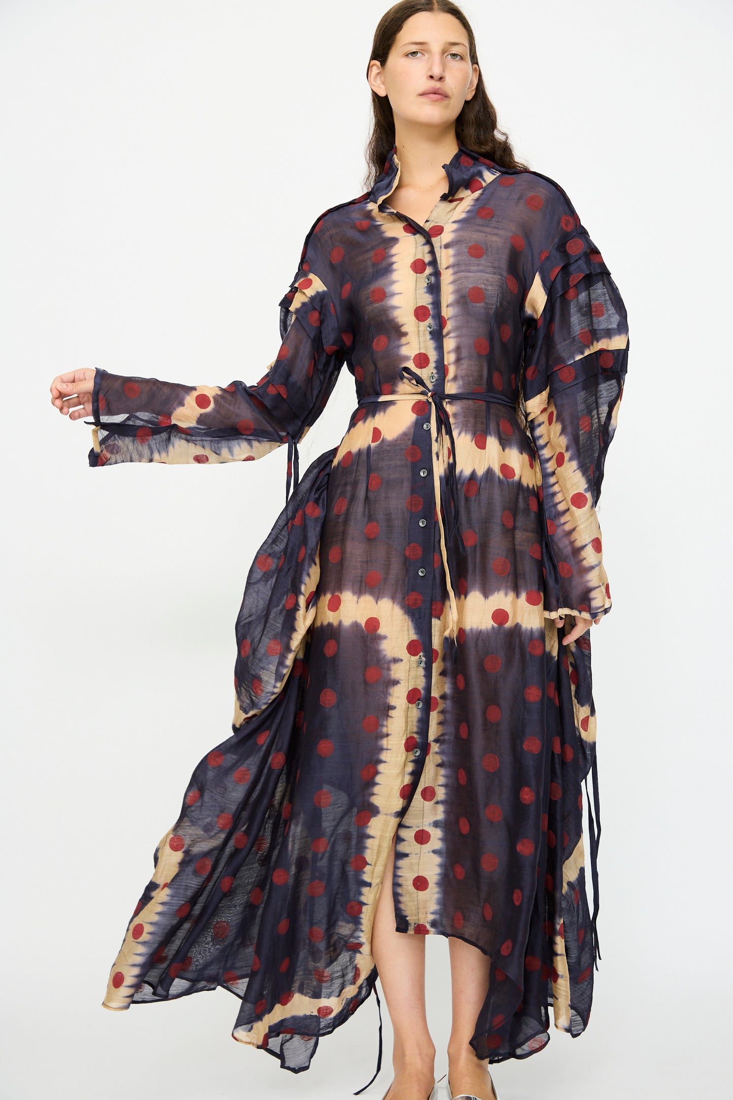 A woman wears the TIGRA TIGRA Silk Chanderi Teatime Dress in navy, cream, and red. This sheer maxi dress features long sleeves, an asymmetrical hem, front buttons, and a mix of polka dots with tie-dye patterns.