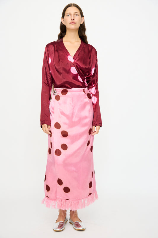 A woman stands against a white background wearing the TIGRA TIGRA Silk Mashroo Moon Dye Wrap Skirt in Pink, styled with a maroon satin top and silver shoes.