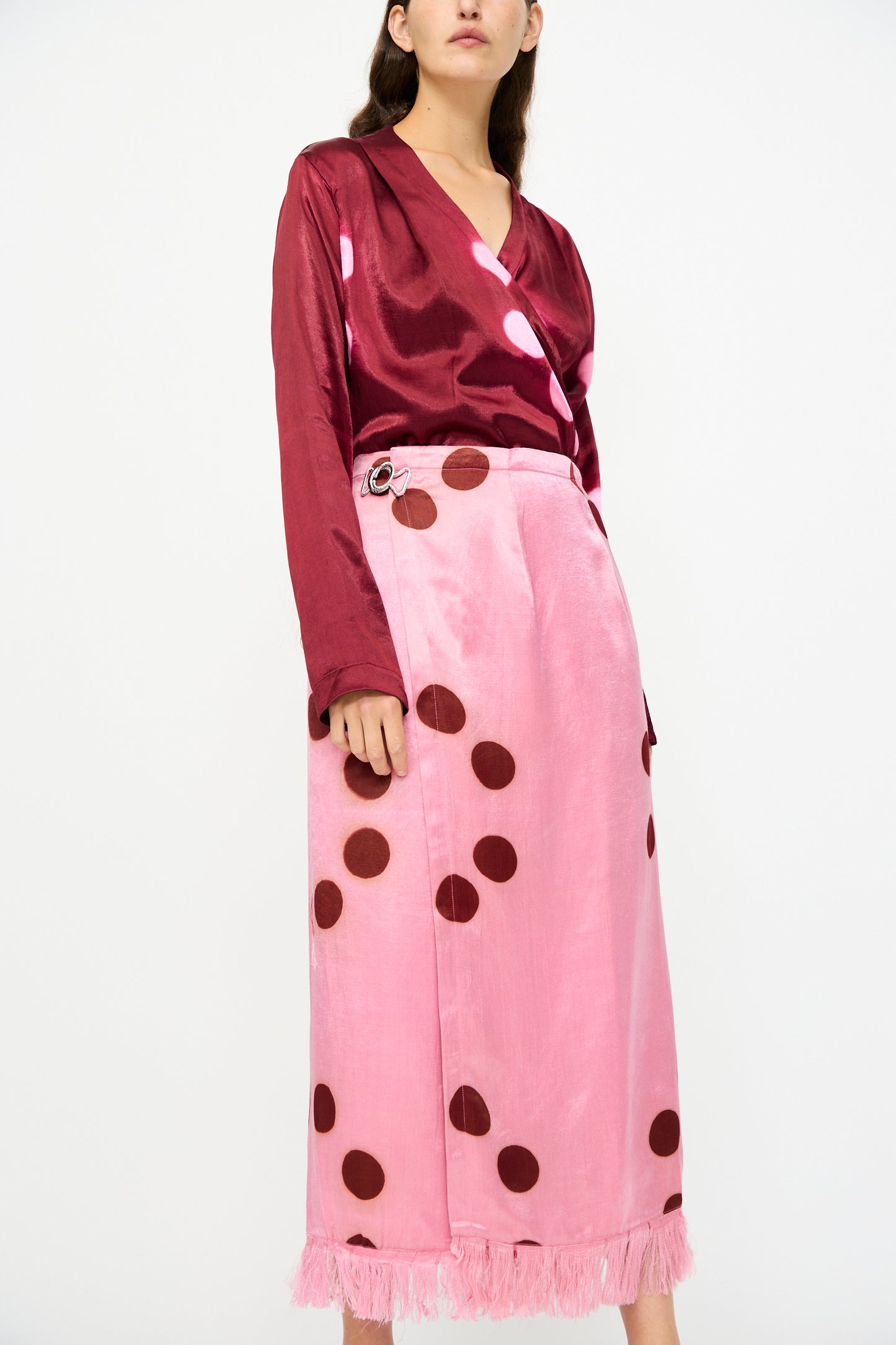 A woman stands against a plain white background wearing a shiny burgundy long-sleeve top and the TIGRA TIGRA Silk Mashroo Moon Dye Wrap Skirt in Pink.