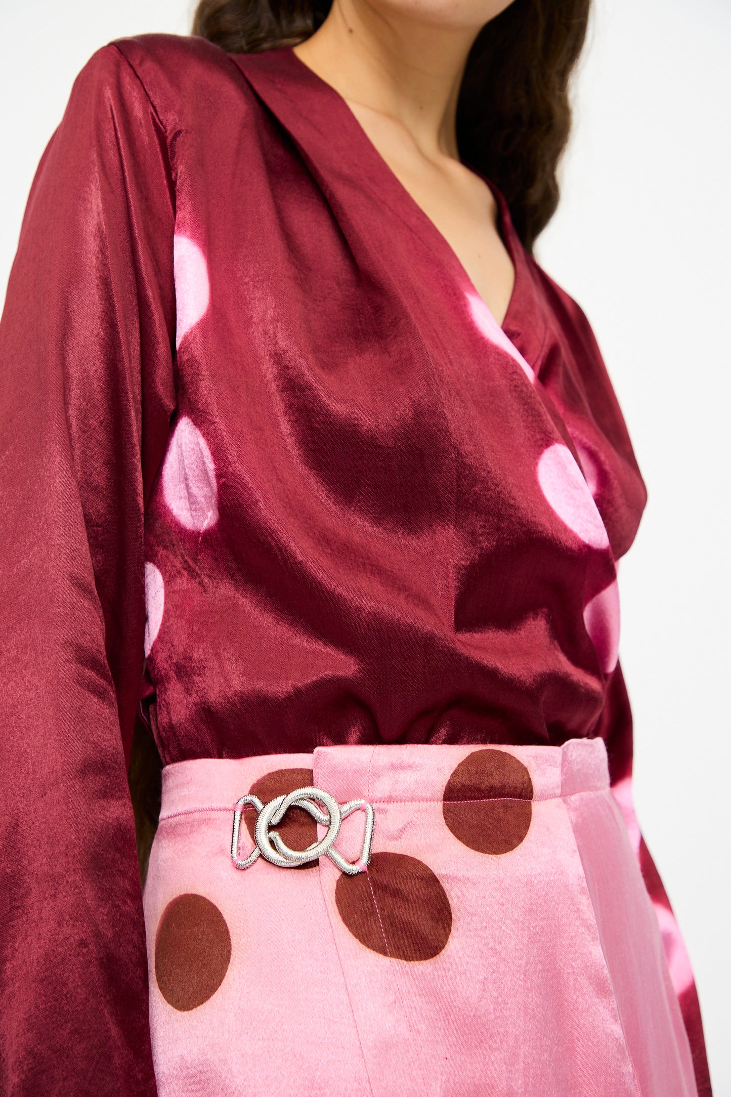 A person wearing a shiny maroon blouse with pink polka dots, light pink pants with brown polka dots, a silver belt buckle, and the TIGRA TIGRA Silk Mashroo Moon Dye Wrap Skirt in Pink.
