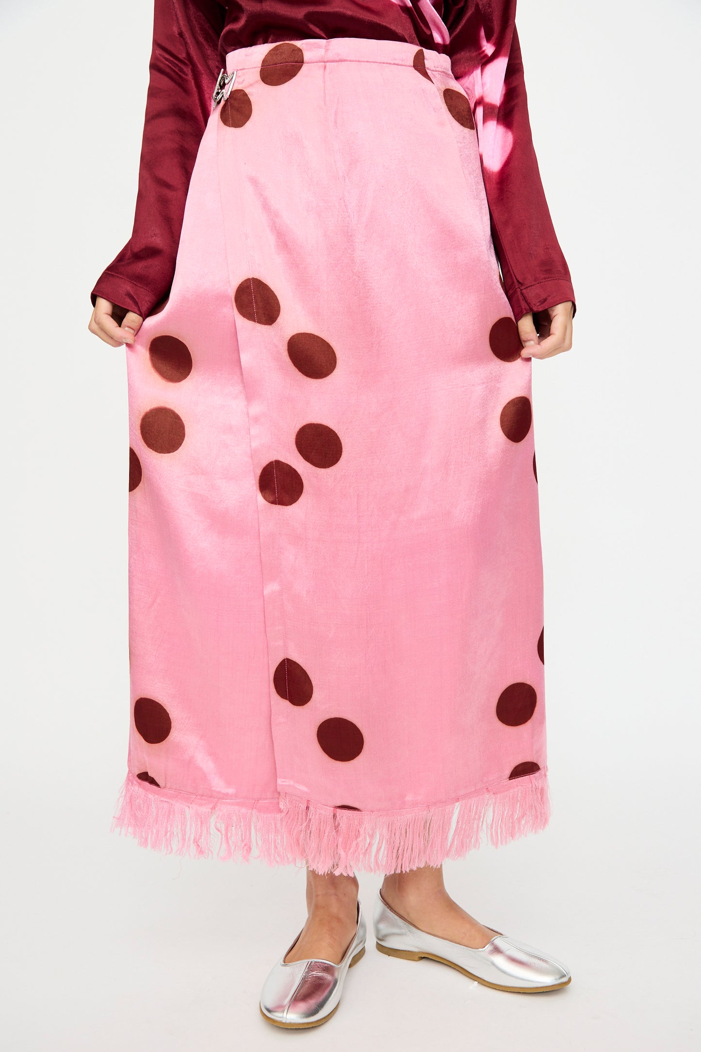 A person wearing the TIGRA TIGRA Silk Mashroo Moon Dye Wrap Skirt in Pink, a shiny burgundy top, and silver flat shoes.