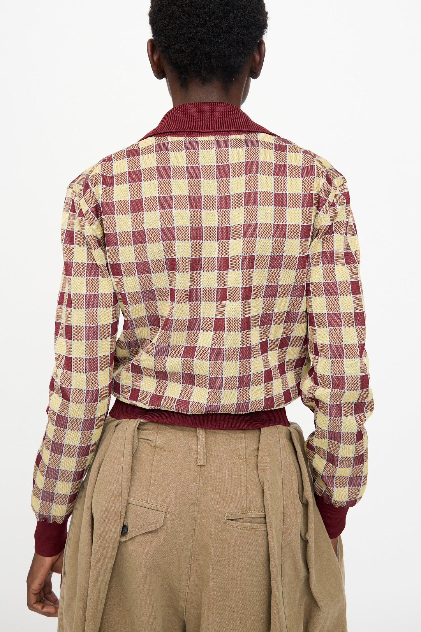 A person is seen from behind wearing the TOGA ARCHIVES Check Knit Cardigan in Dark Red and tan high-waisted pants with belt loops and pleats, standing against a plain white background.