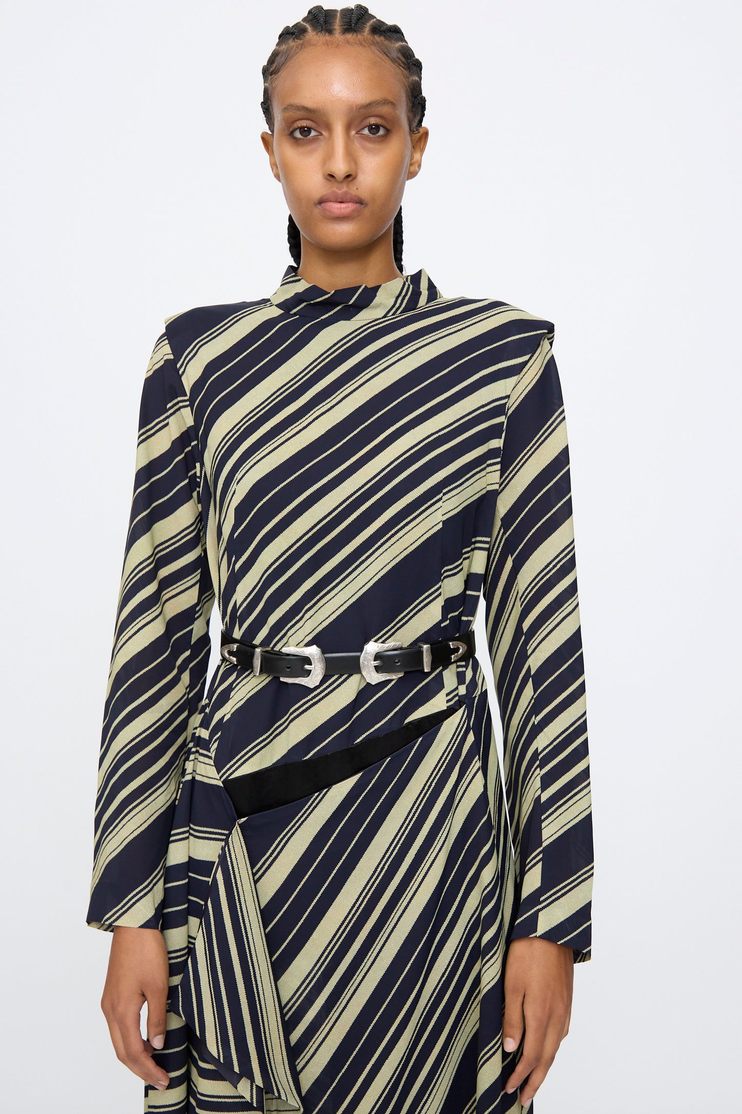 A person stands against a plain light background wearing a long-sleeve black and beige striped dress, styled with the TOGA ARCHIVES Double Buckle Belt in Black featuring silver buckles at the waist.