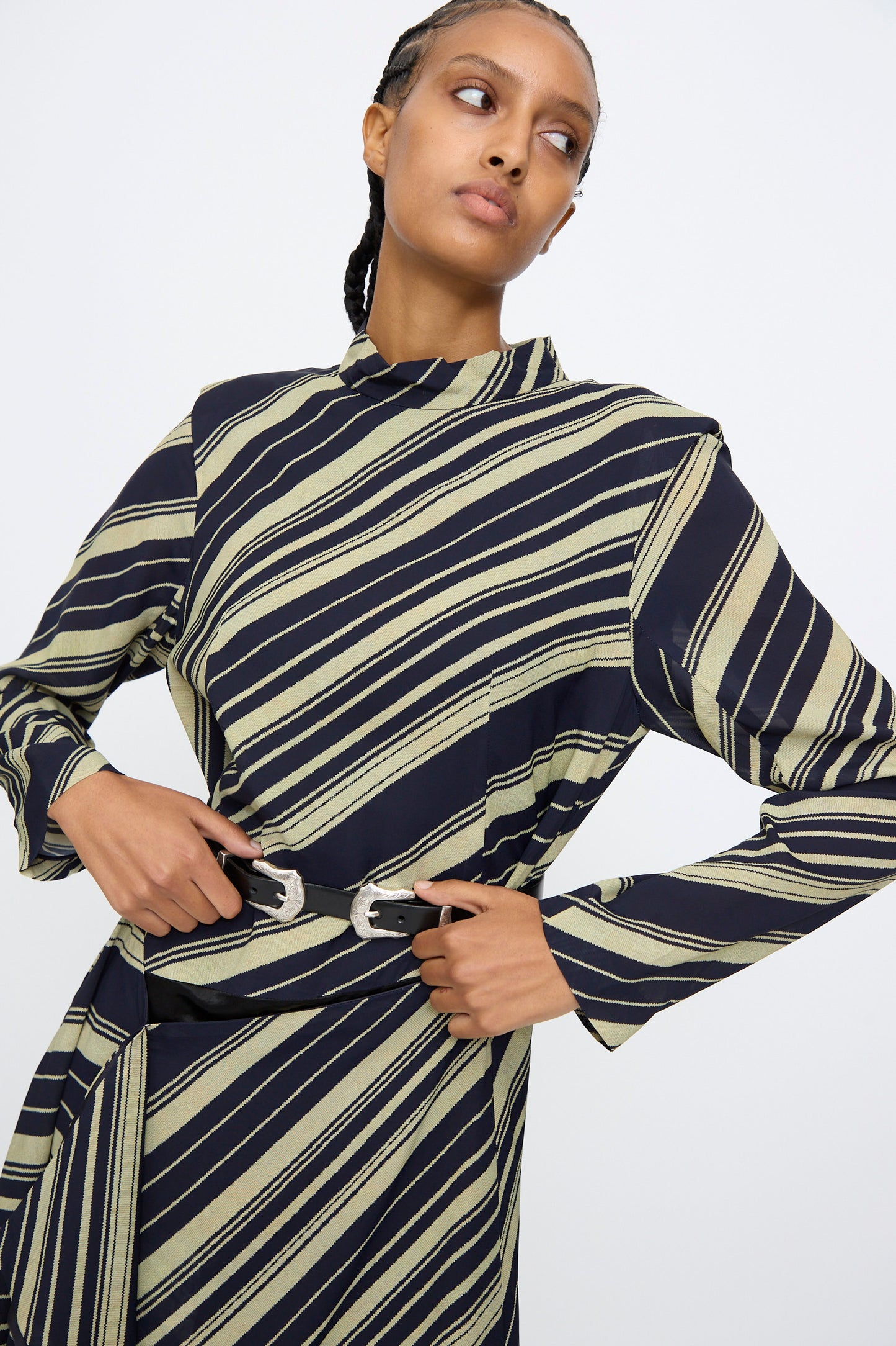 A person wearing a long-sleeved dress with diagonal black and beige stripes pairs it with the TOGA ARCHIVES Double Buckle Belt in Black, featuring silver buckles, and stands against a plain light background.