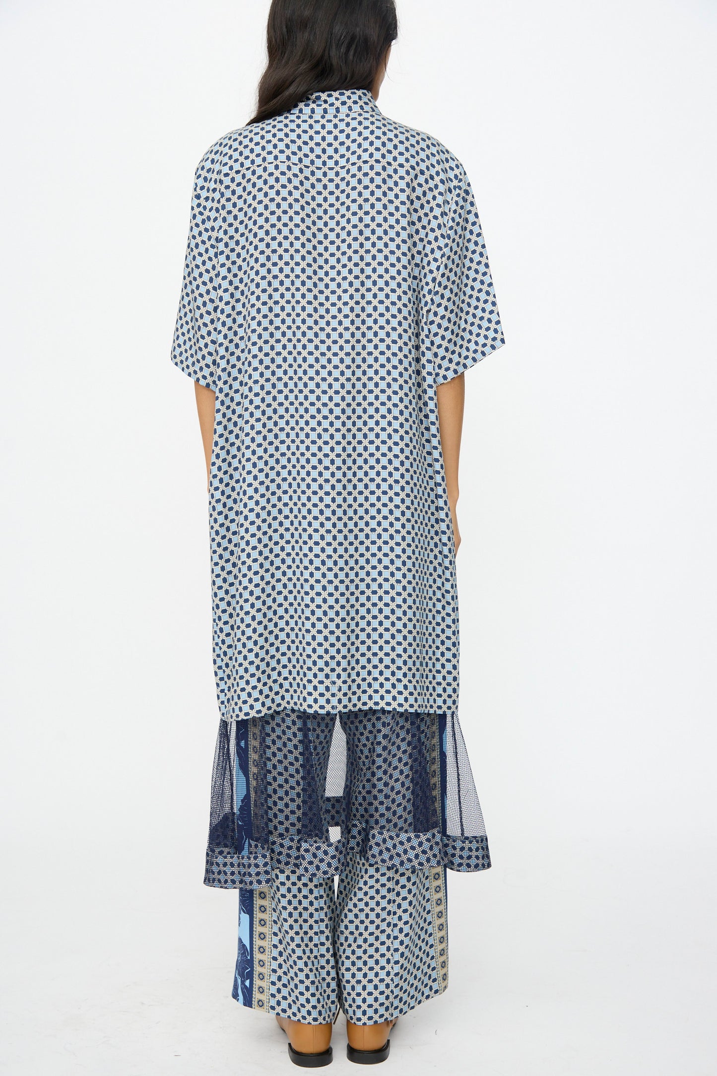 Person standing with back to camera, wearing the TOGA ARCHIVES Inner Print Dress in Light Blue, a loose cupra dress featuring blue and white patterns and sheer lower panels, set against a plain white background.