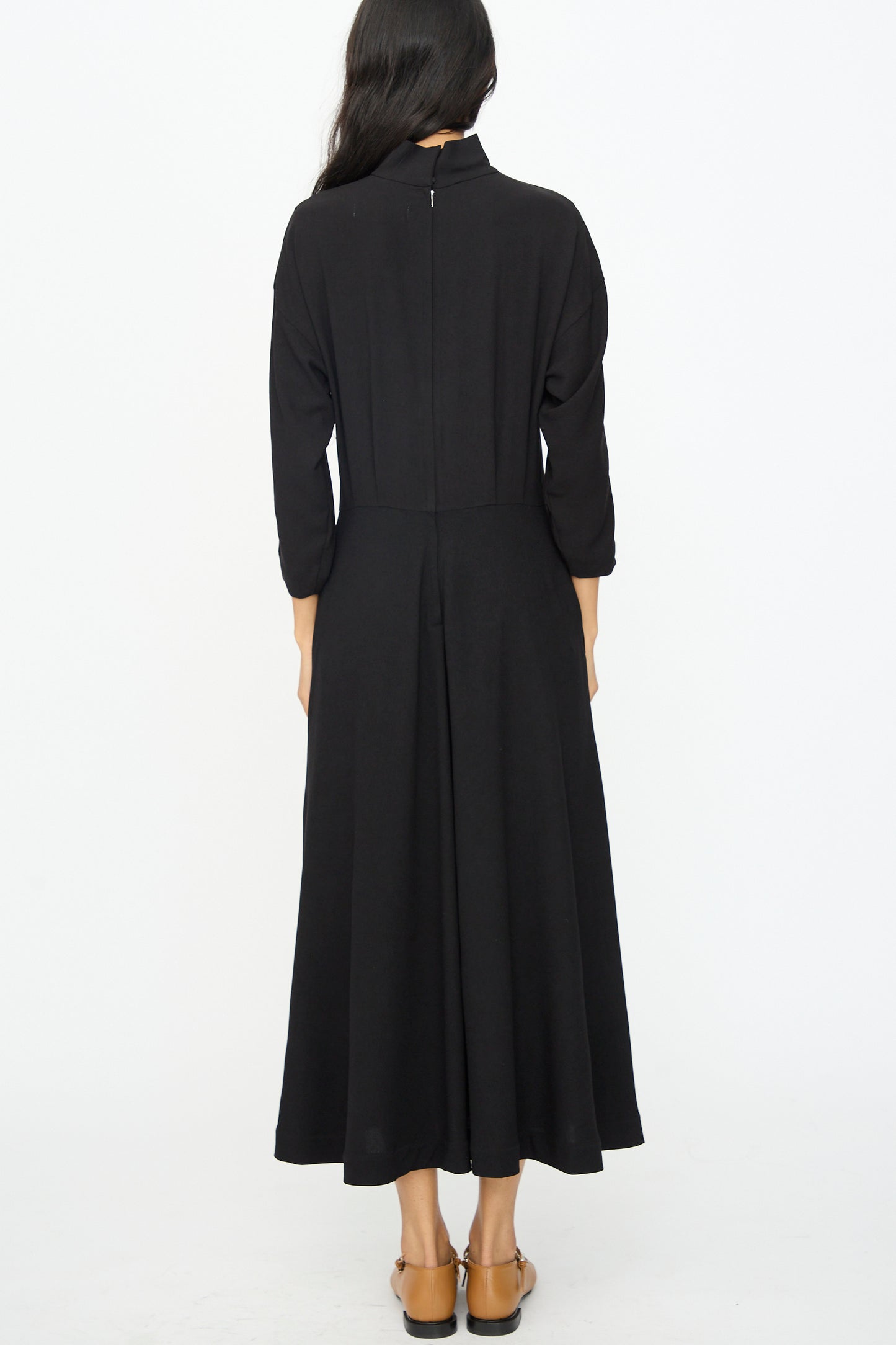 A woman with long dark hair stands with her back to the camera against a white background, wearing the TOGA ARCHIVES Lace Dress in Black and brown flats.