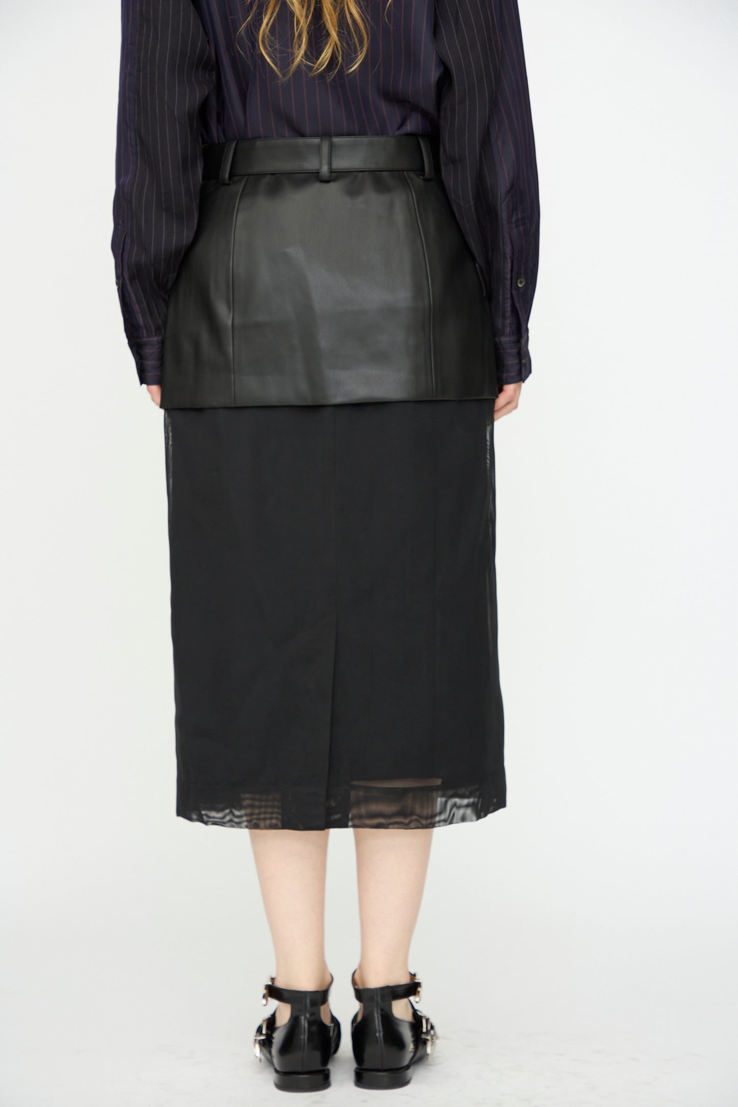 Person facing away, wearing a black pinstripe shirt, TOGA ARCHIVES Leather Mesh Skirt in Black layered over a black slip skirt, and black buckle sandals, set against a plain white background.