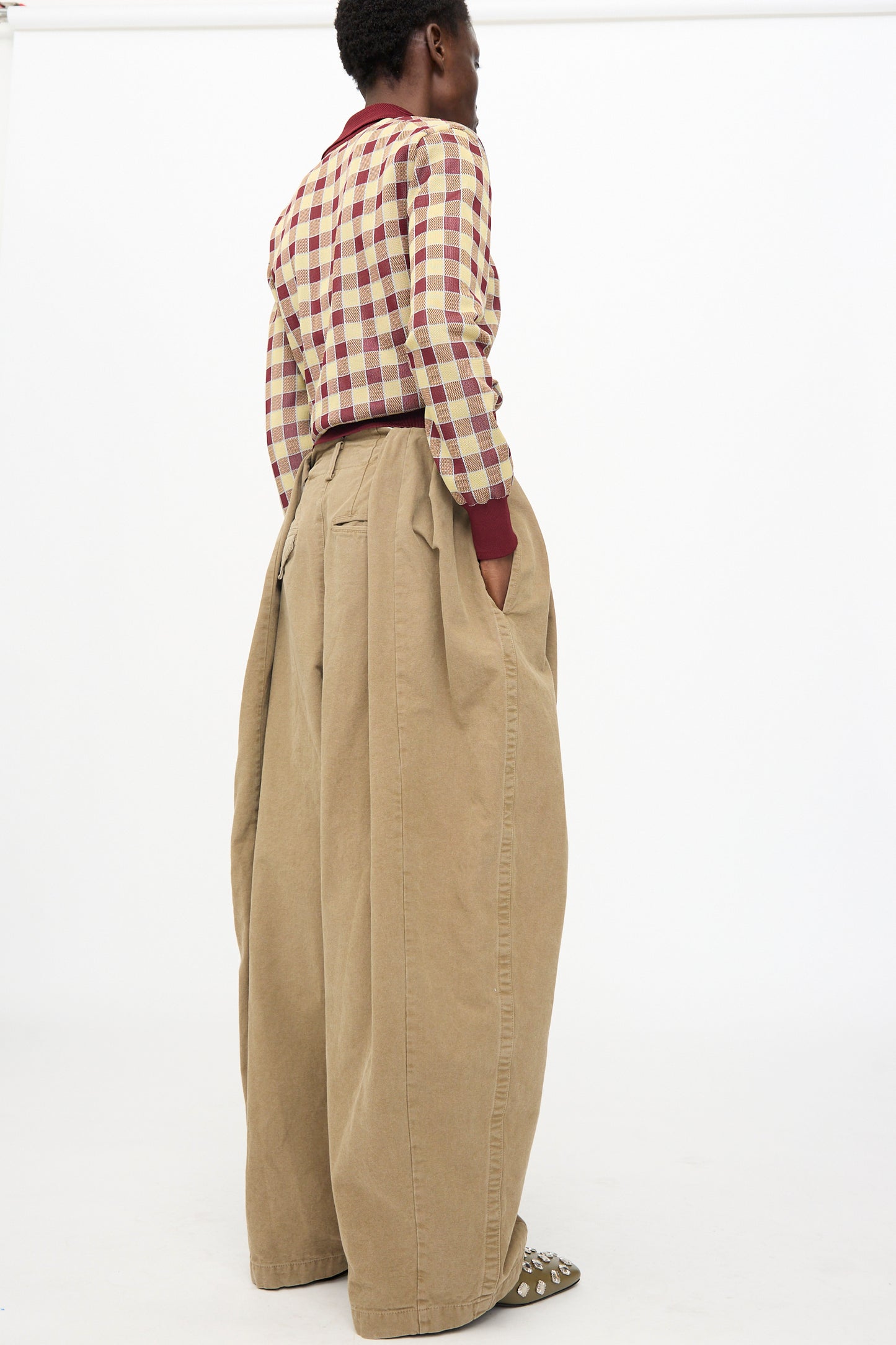 A person stands sideways against a white background, wearing TOGA ARCHIVES’ Oxford Wide Pants in Beige and a red and white checkered shirt, hands in pockets.