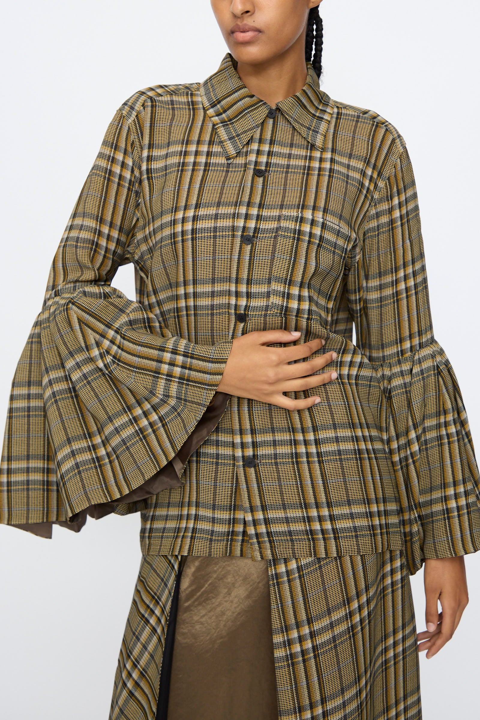 A person wears the TOGA ARCHIVES Rayon Print Shirt in Brown, featuring a brown and yellow plaid pattern with wide flared sleeves, paired with a matching skirt, standing against a plain white background. This stylish shirt is made in Japan.