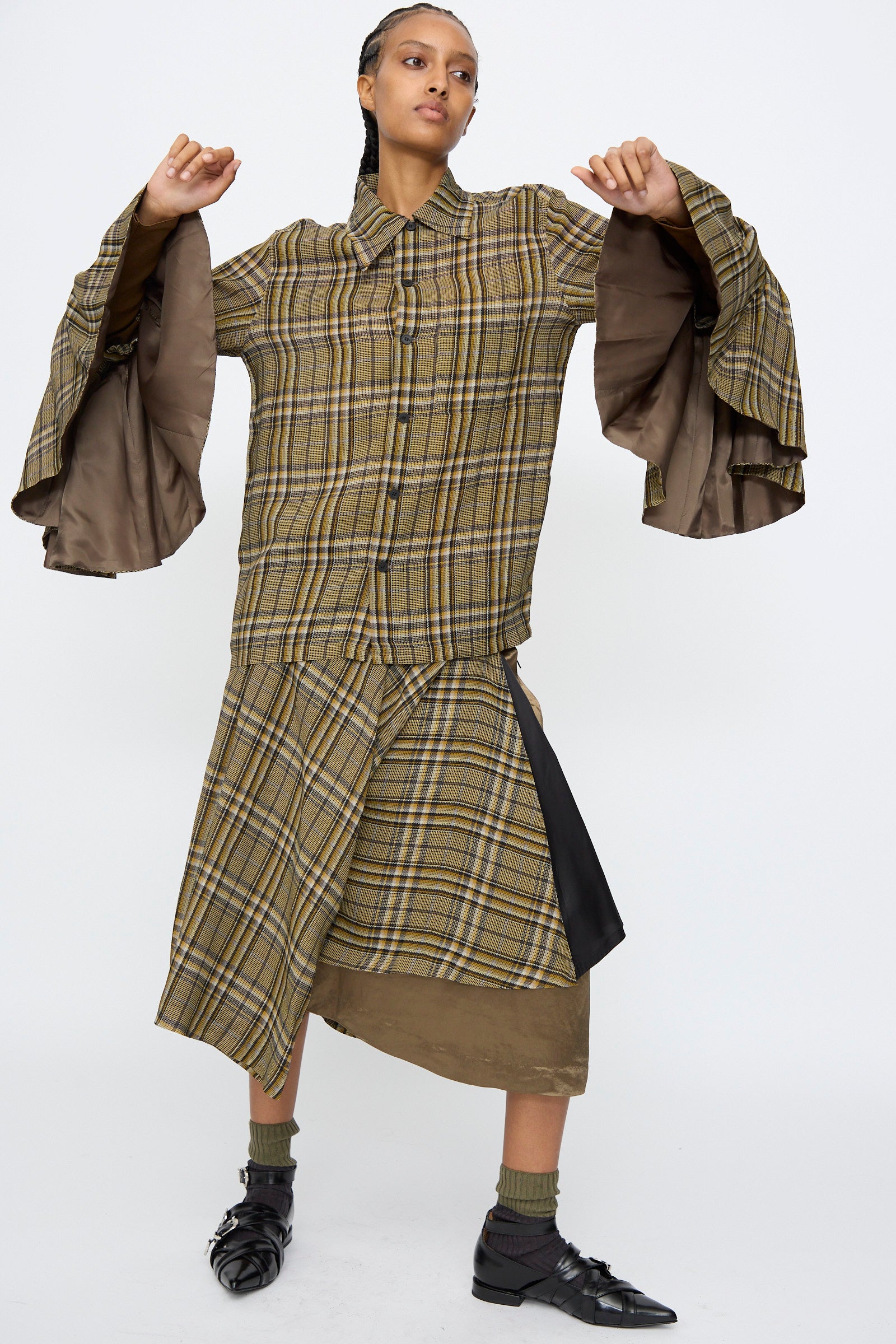 A person stands with arms raised, wearing the TOGA ARCHIVES Rayon Print Shirt in Brown. The classic design, made in Japan, is paired with a matching skirt, dark socks, and black shoes against a plain white background.