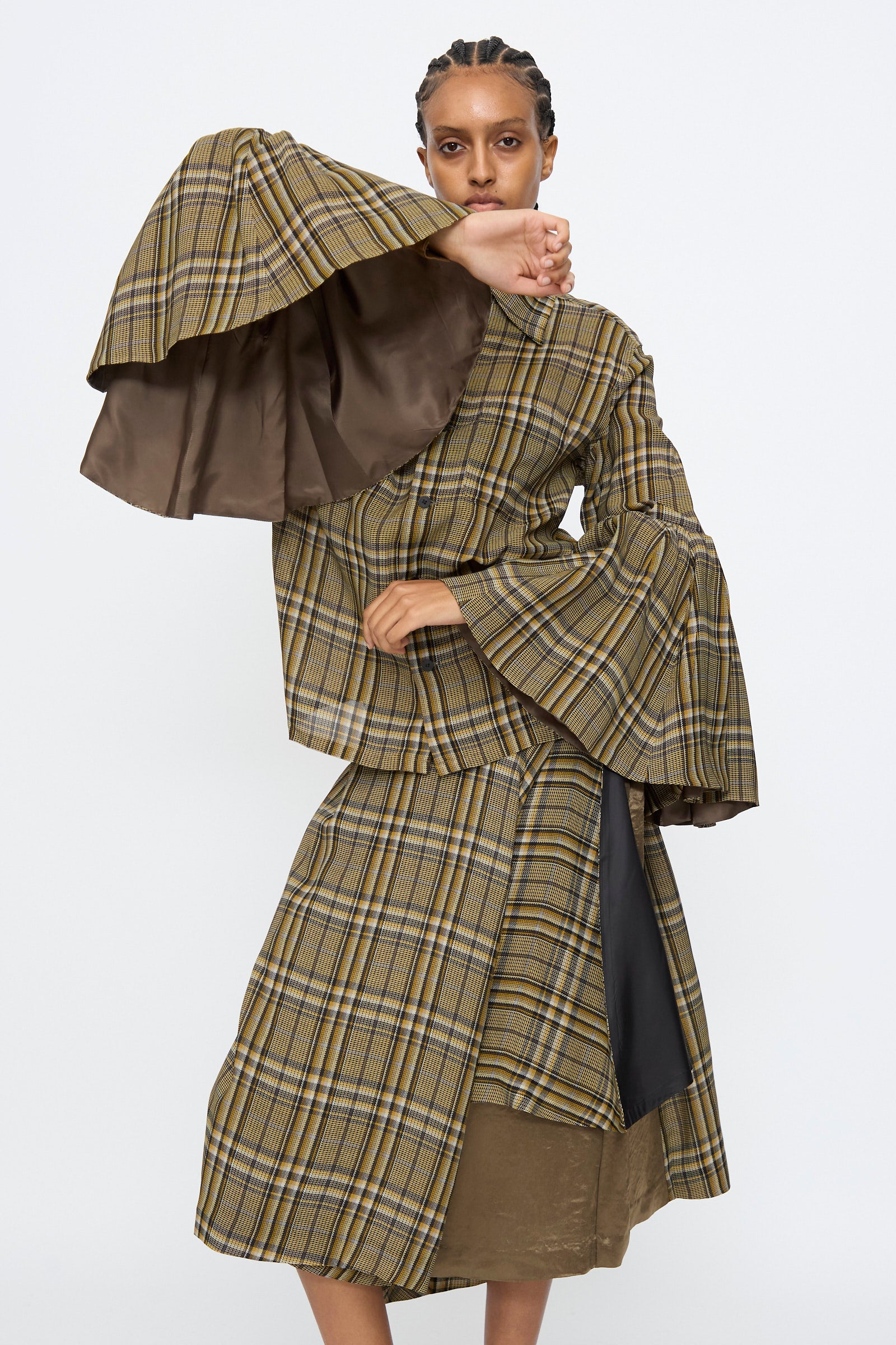 A person models the TOGA ARCHIVES Rayon Print Shirt in Brown, featuring wide flared sleeves and a plaid pattern, while posing against a plain white background with one arm partly covering their face.