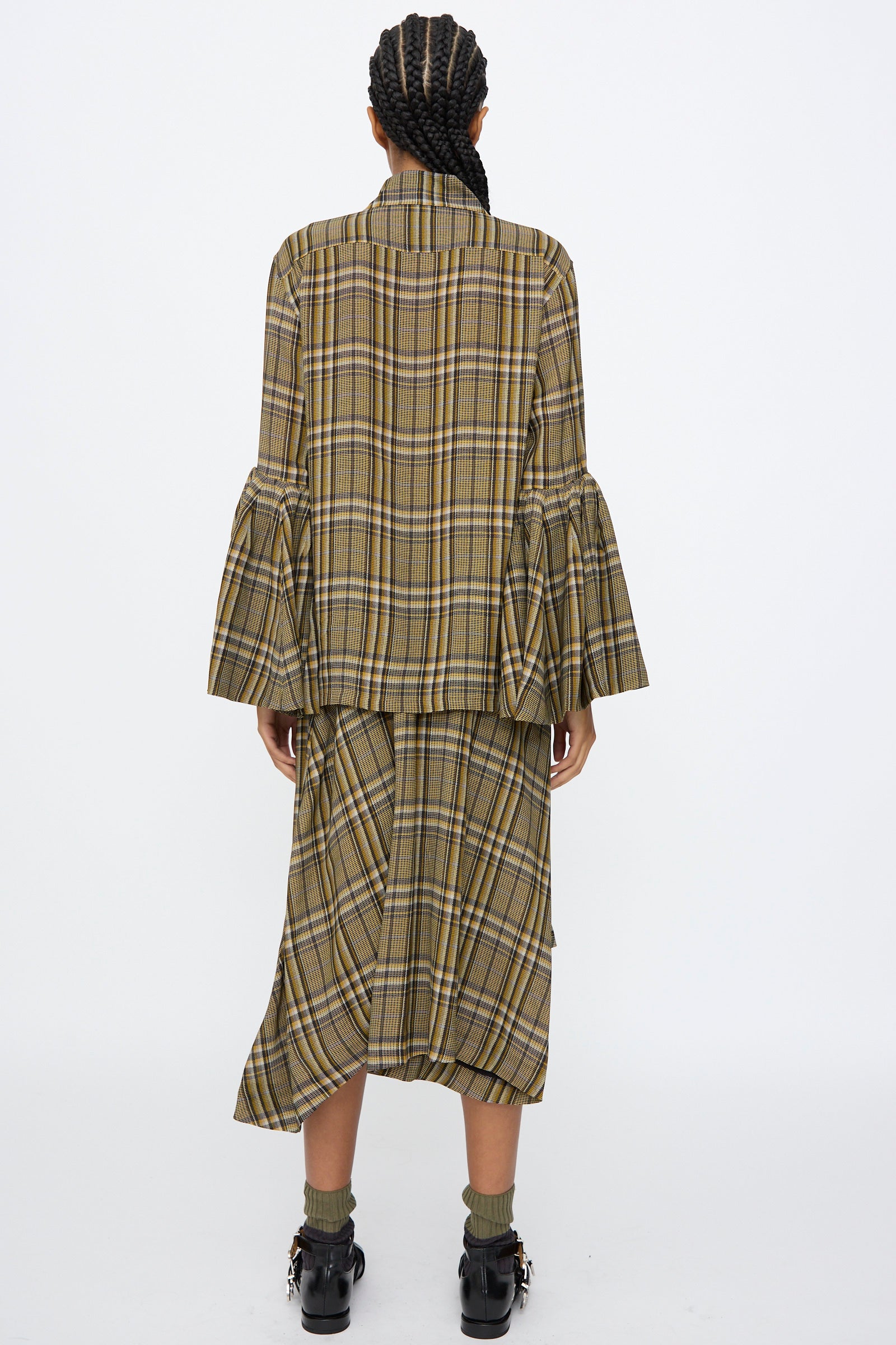 A person with braided hair is shown from the back wearing the TOGA ARCHIVES Rayon Print Shirt in Brown as a loose jacket with flared sleeves, paired with a matching below-the-knee skirt, ankle socks, and black shoes.