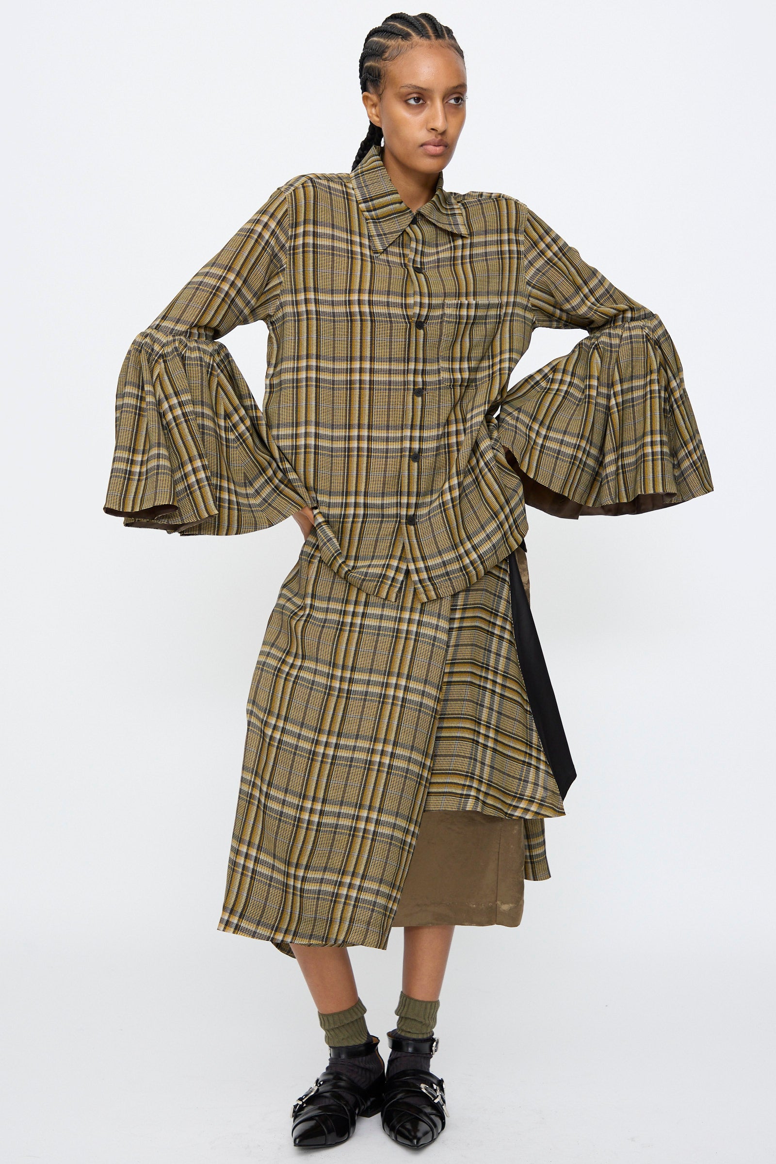 A person wearing the TOGA ARCHIVES Rayon Print Shirt in Brown, featuring a plaid design with flared sleeves, paired with a matching skirt and brown underlayer, standing against a plain white background.