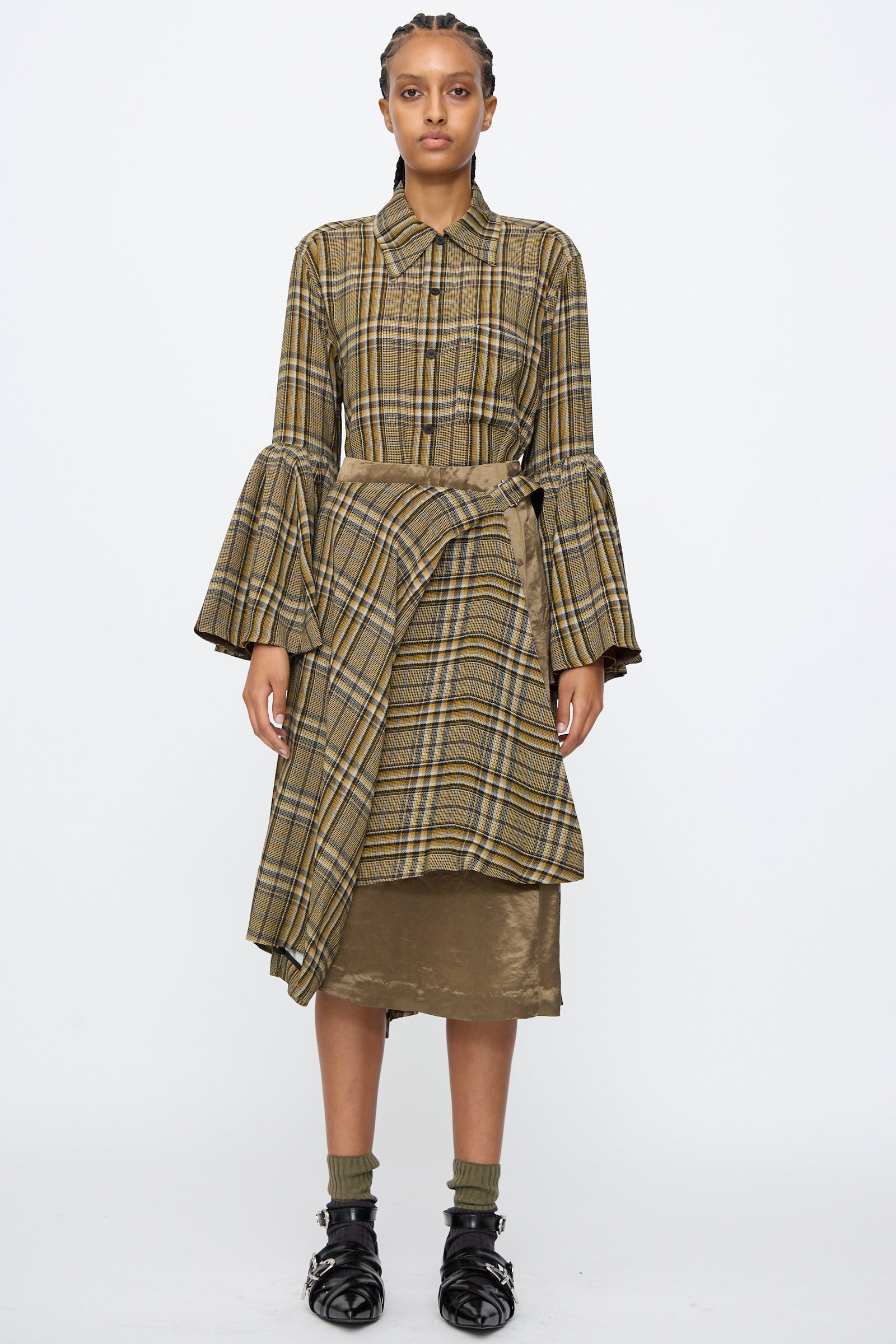 A person stands facing forward, wearing the TOGA ARCHIVES Rayon Print Skirt in Brown, layered over a metallic brown asymmetrical midi skirt, paired with a tan and brown plaid shirt, socks, and black sandals.