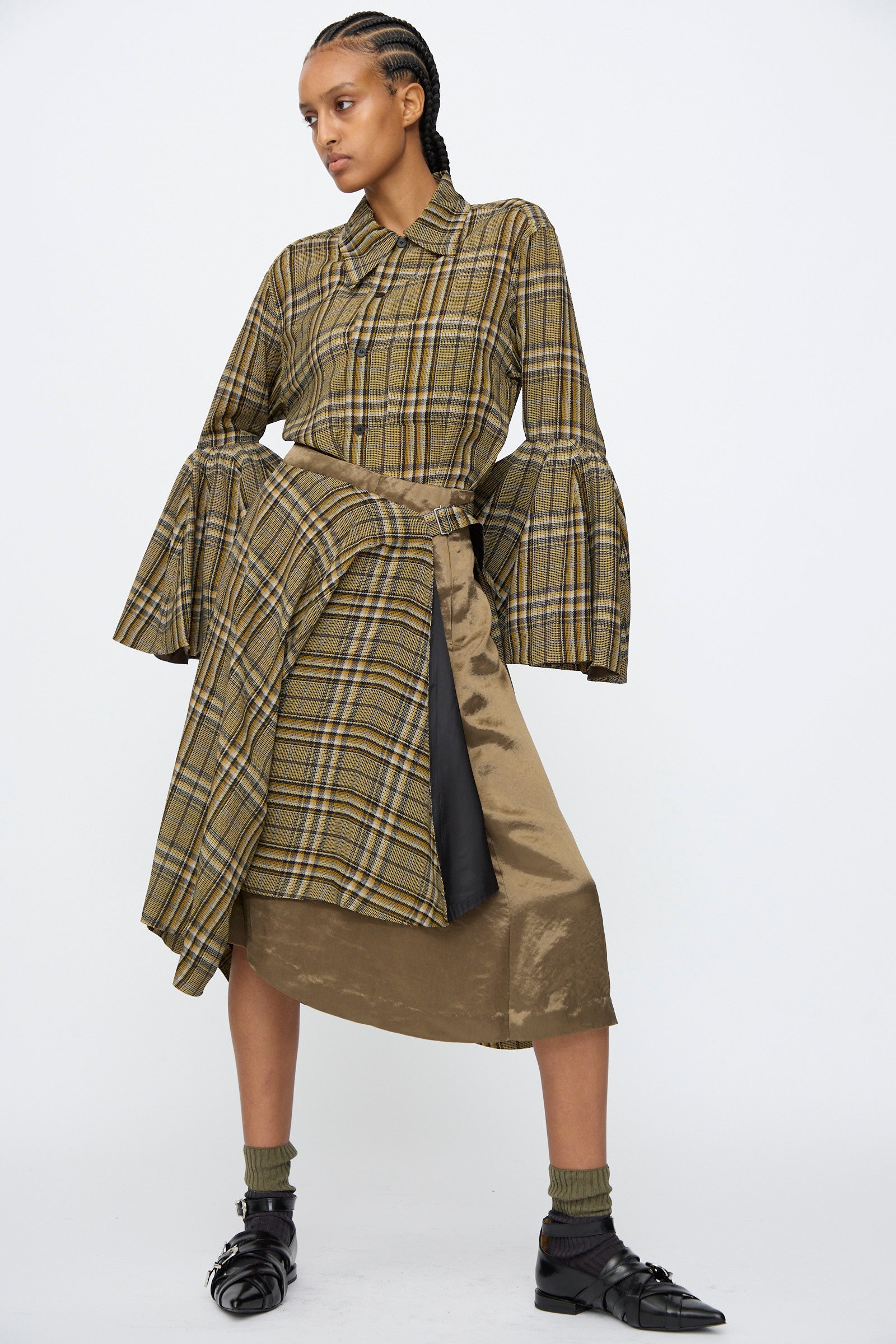 A model wears the Rayon Print Skirt in Brown by TOGA ARCHIVES, styled with a plaid top, gold skirt layered beneath, olive socks, and black shoes, posing against a plain white background.