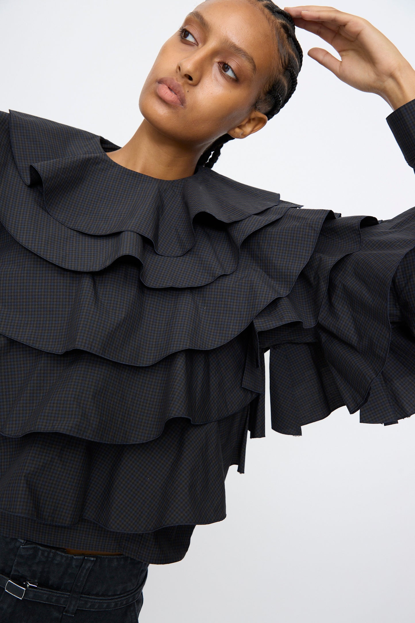 A person poses with one arm raised against a plain background, wearing the TOGA ARCHIVES Typewriter Check Frill Blouse in Navy, featuring ruffled layers, a boxy fit, and wide long sleeves.