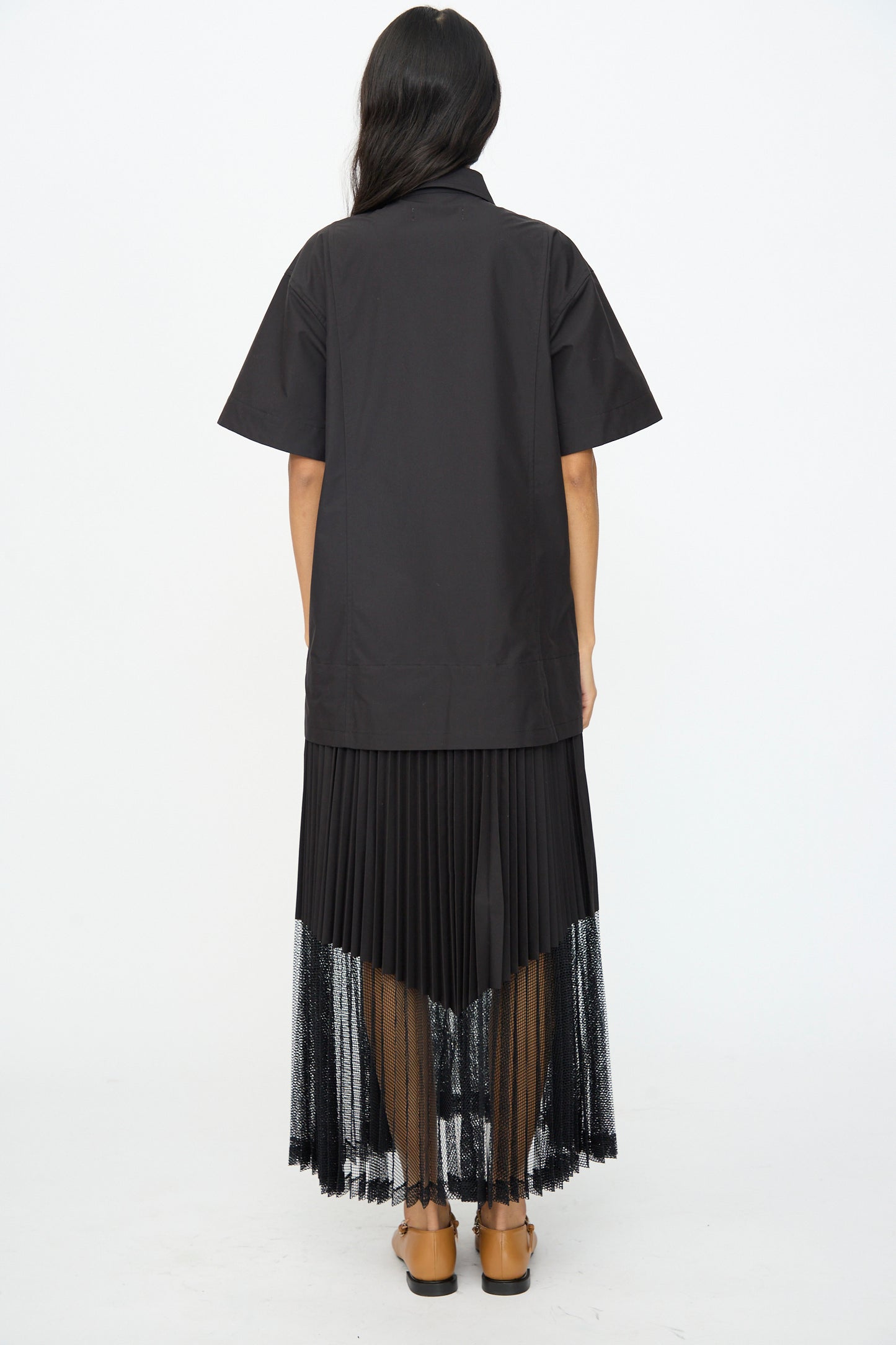 A person faces away, wearing the TOGA ARCHIVES Typewriter Mesh Dress in Black, featuring short sleeves and pleated details, paired with tan flat shoes.