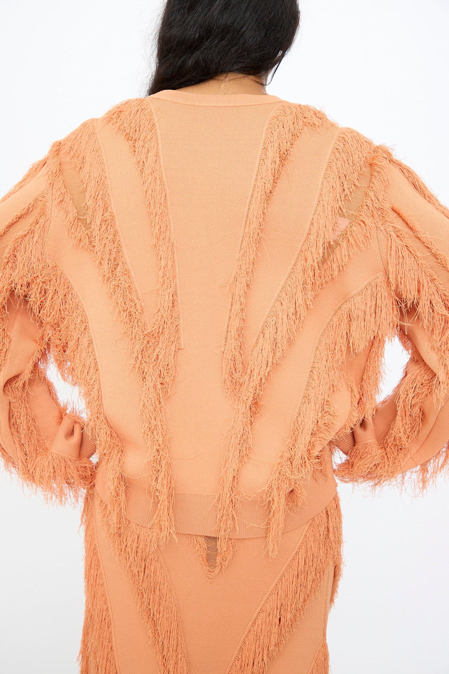 A person with long dark hair is shown from the back, wearing the Ulla Johnson Aubree Knit Pullover in Tulip, a peach-colored jacquard sweater with textured, fringed accents on the sleeves and back.