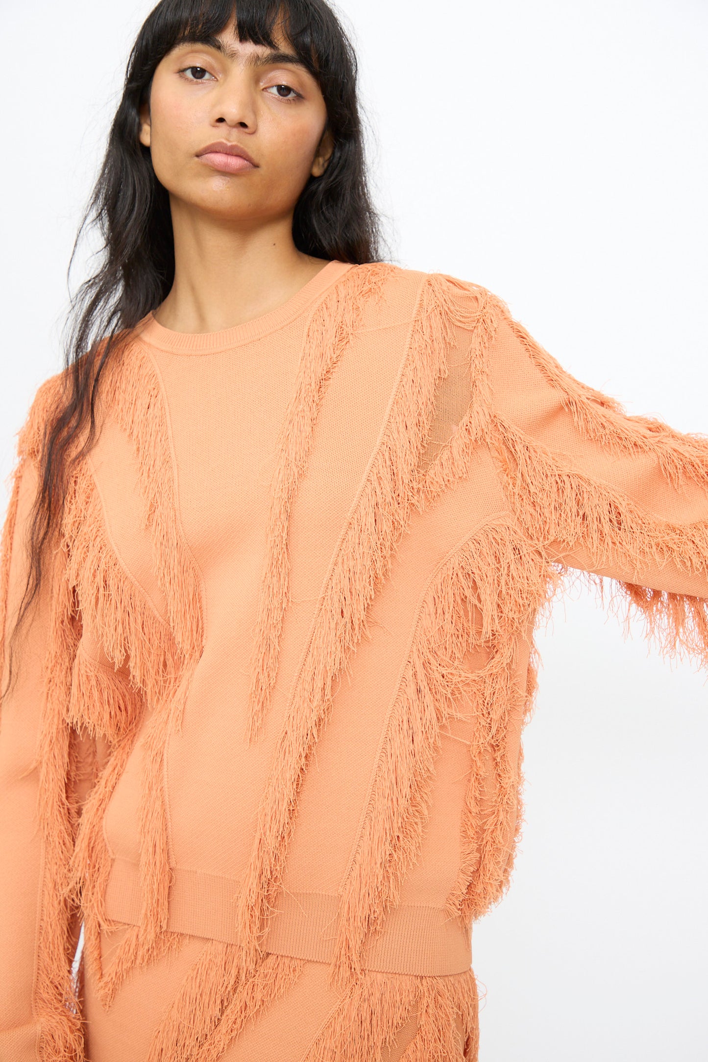 A person wearing the Ulla Johnson Aubree Knit Pullover in Tulip, featuring textured fringe, stands against a plain white background.