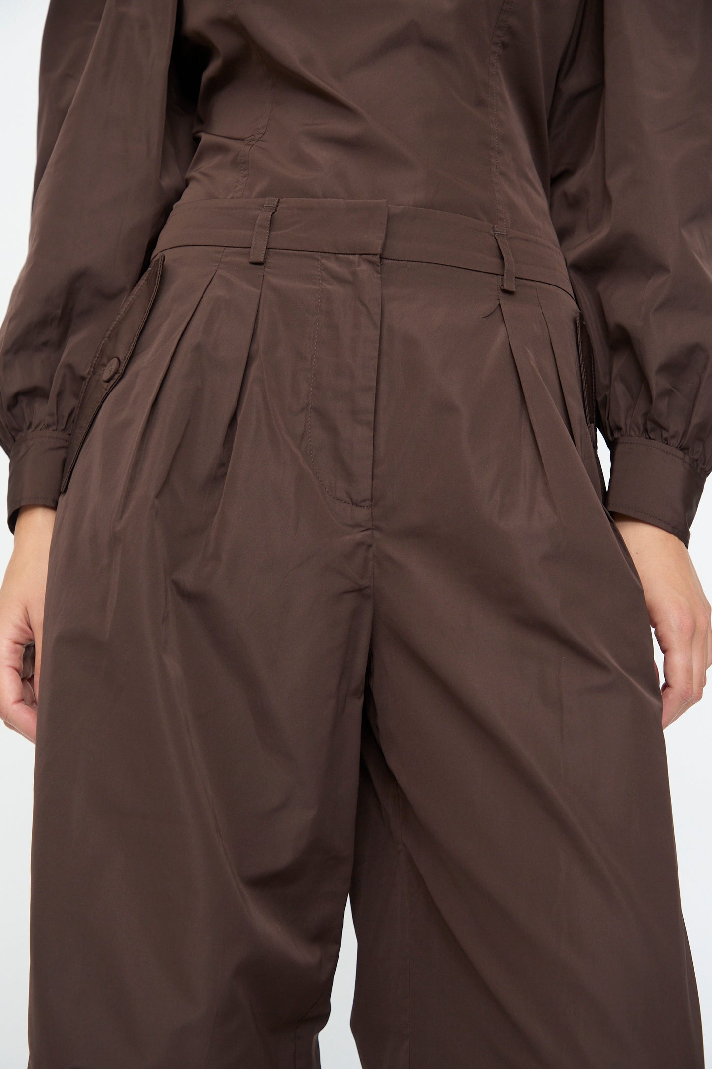 Close-up of a person wearing Ulla Johnson’s Charlie Silk Pant in Sequoia, high-waisted with wide legs and pleats, paired with a matching brown top, both crafted from taffeta-finished habotai.