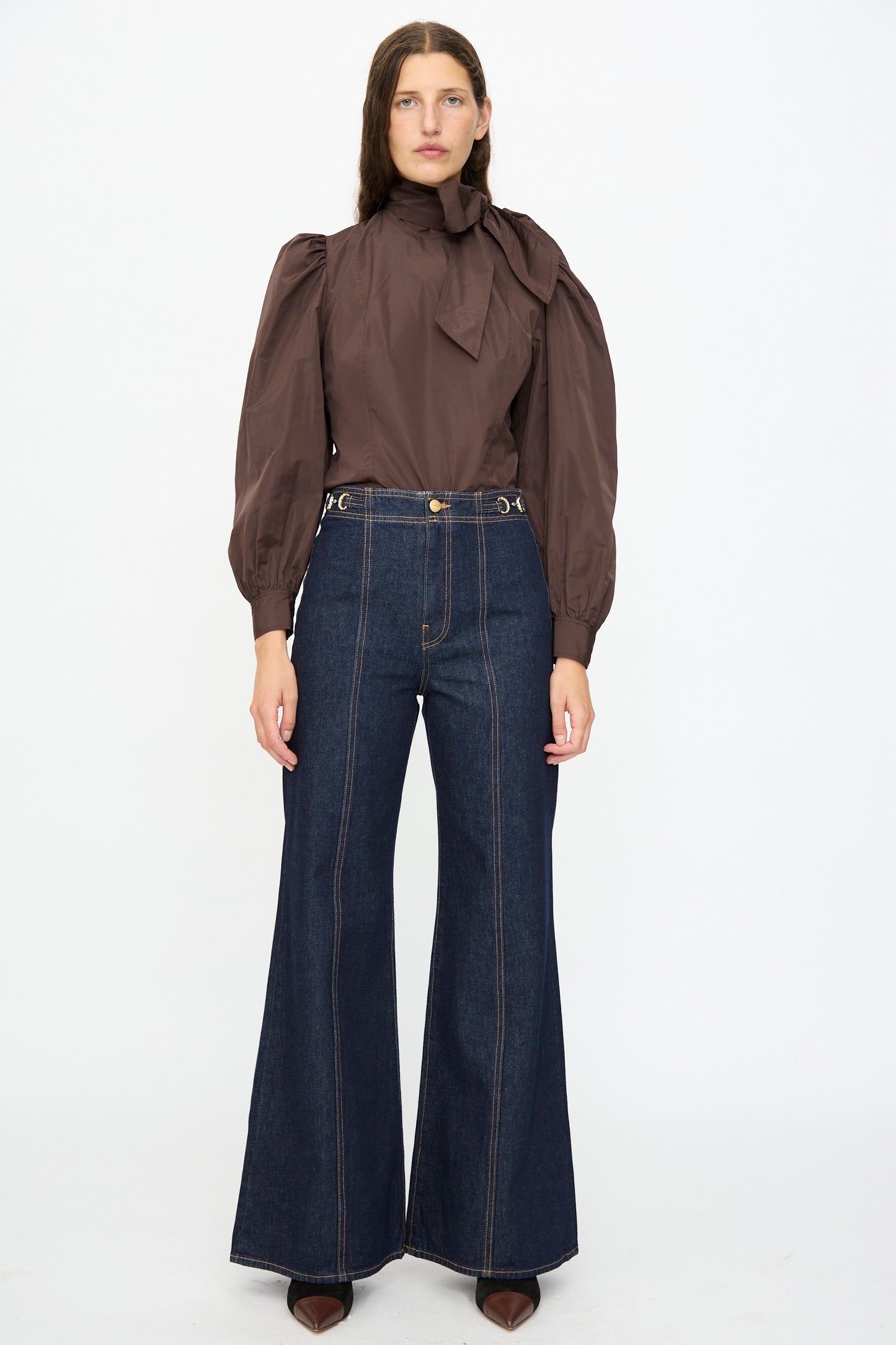 A woman faces forward wearing a brown puff-sleeve blouse with a neck bow and Ulla Johnson’s Delphine High Rise Jean in Tigris, featuring deep indigo cotton and flared legs.