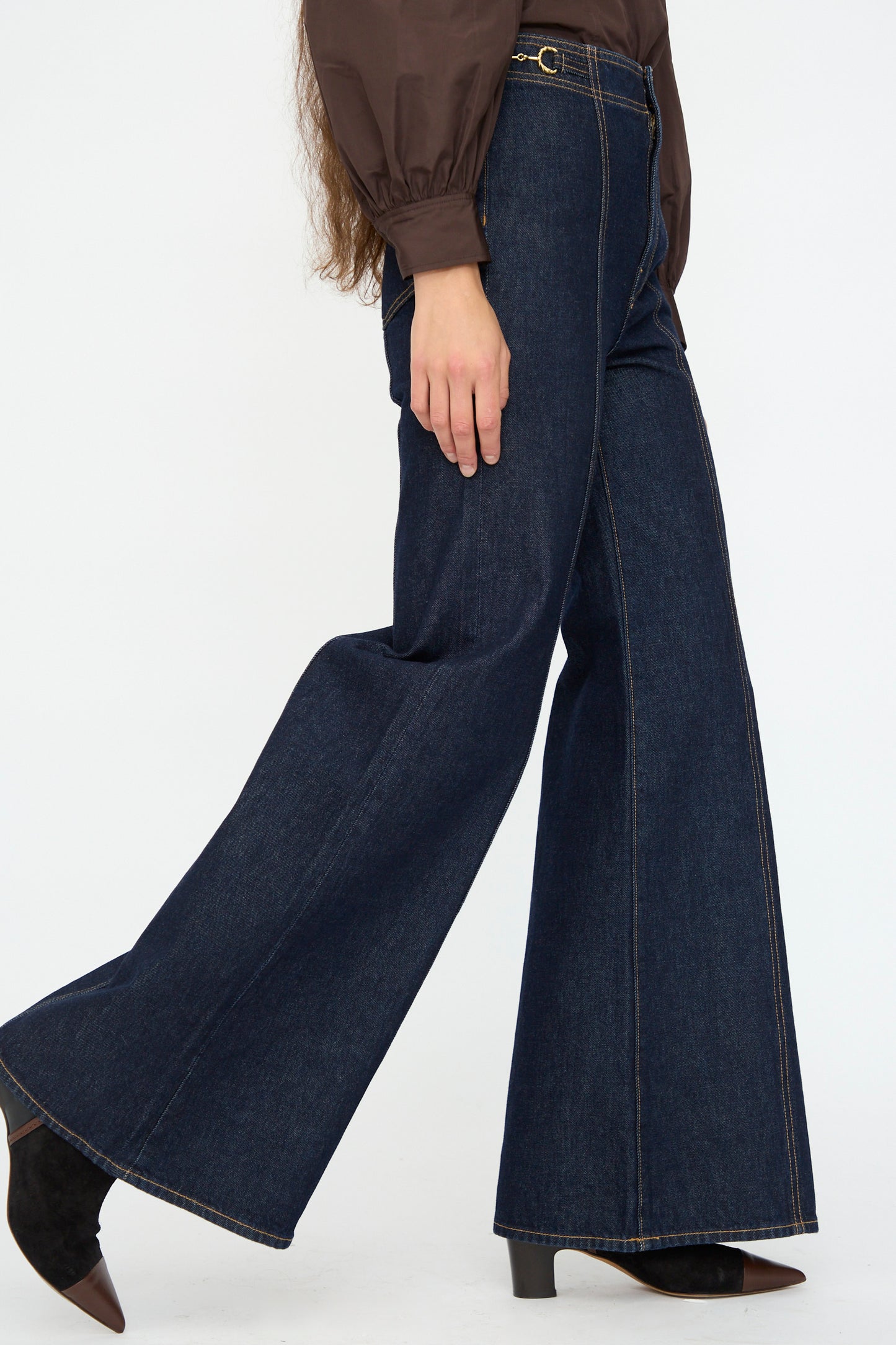 Wearing Ulla Johnson's Delphine High Rise Jean in Tigris, a model walks forward in deep indigo flared jeans and a brown blouse, shown from shoulder down.