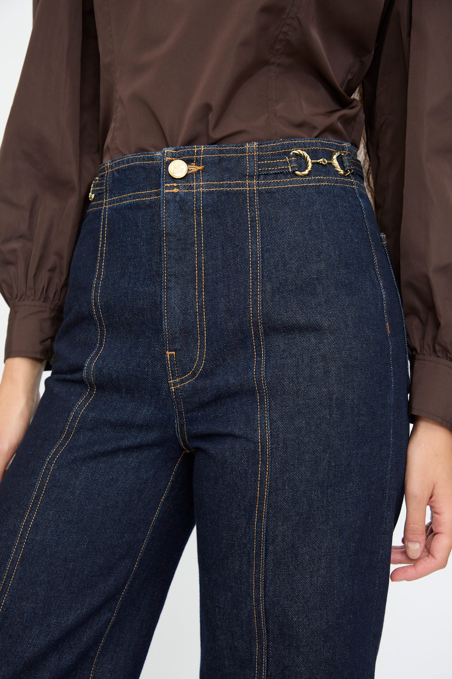 Someone wearing Ulla Johnson’s Delphine High Rise Jean in Tigris—deep indigo, high waisted with a flared leg and contrast stitching—paired with a brown long-sleeve shirt.