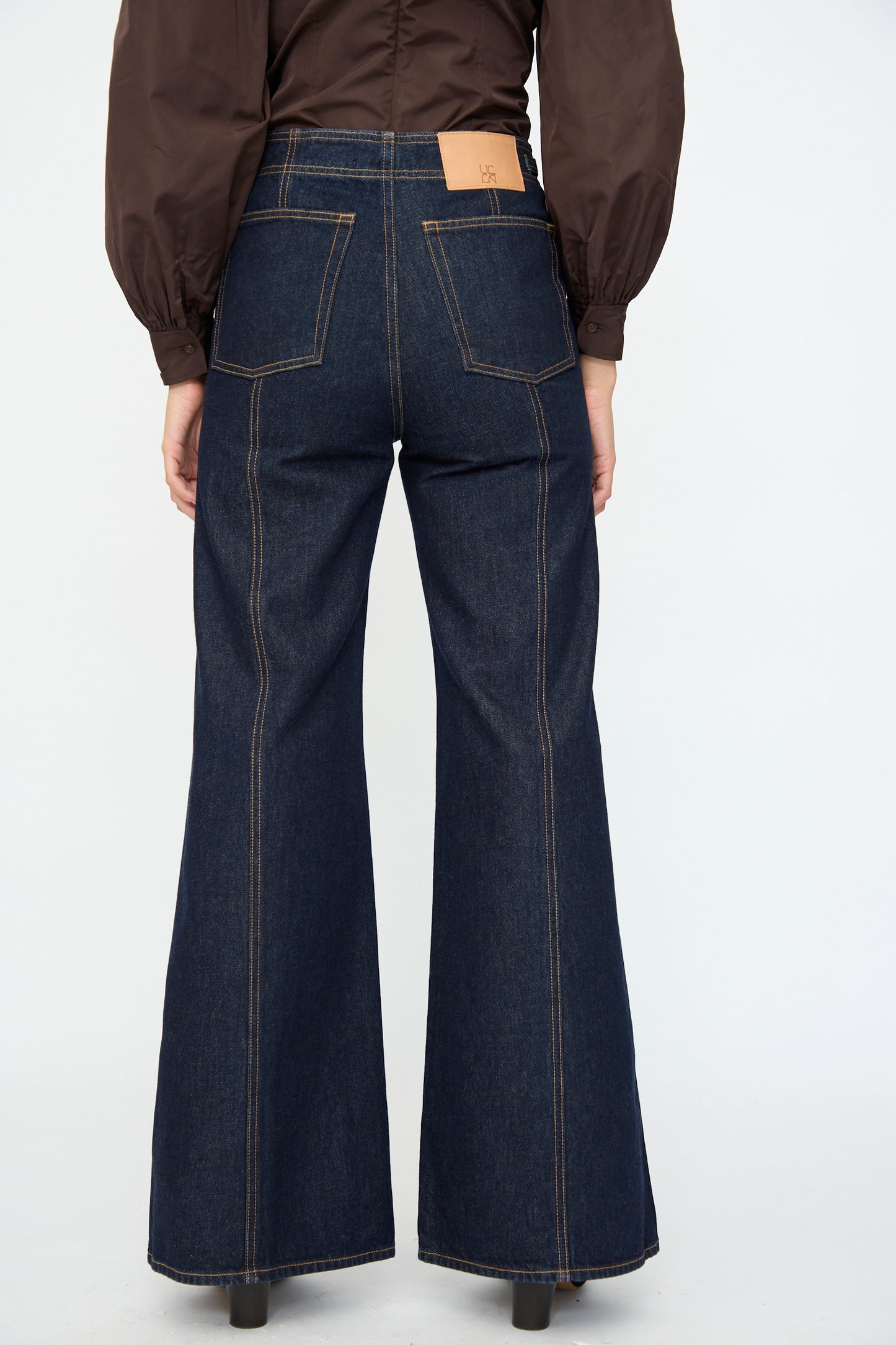 Person shown from the back wearing Ulla Johnson’s Delphine High Rise Jean in Tigris, a deep indigo, high-waisted flared jean, paired with a brown long-sleeve top.