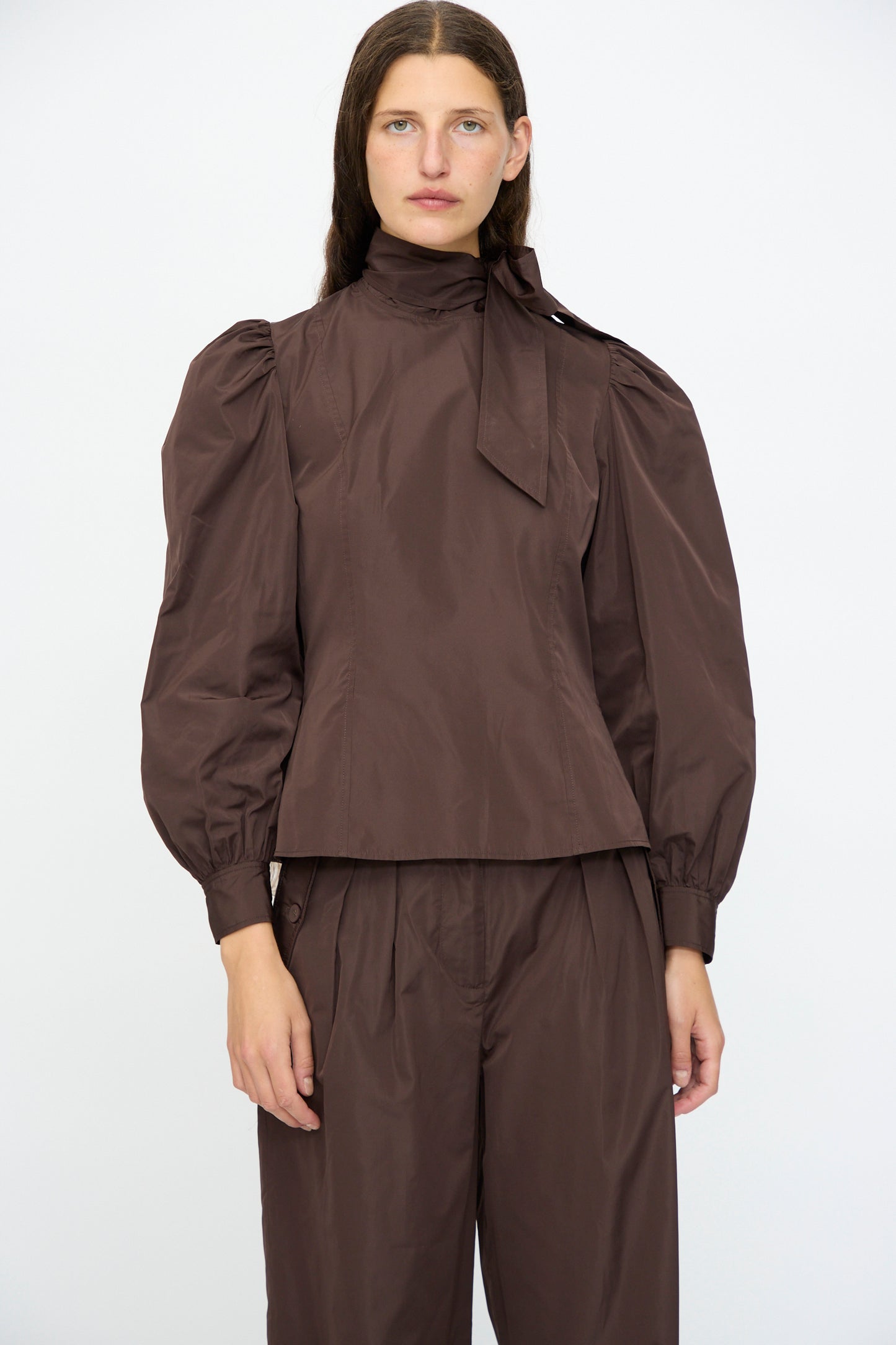 A person stands against a plain background wearing the Ulla Johnson Giorgia Silk Blouse in Sequoia, featuring balloon sleeves and a bow neck, paired with matching brown trousers.