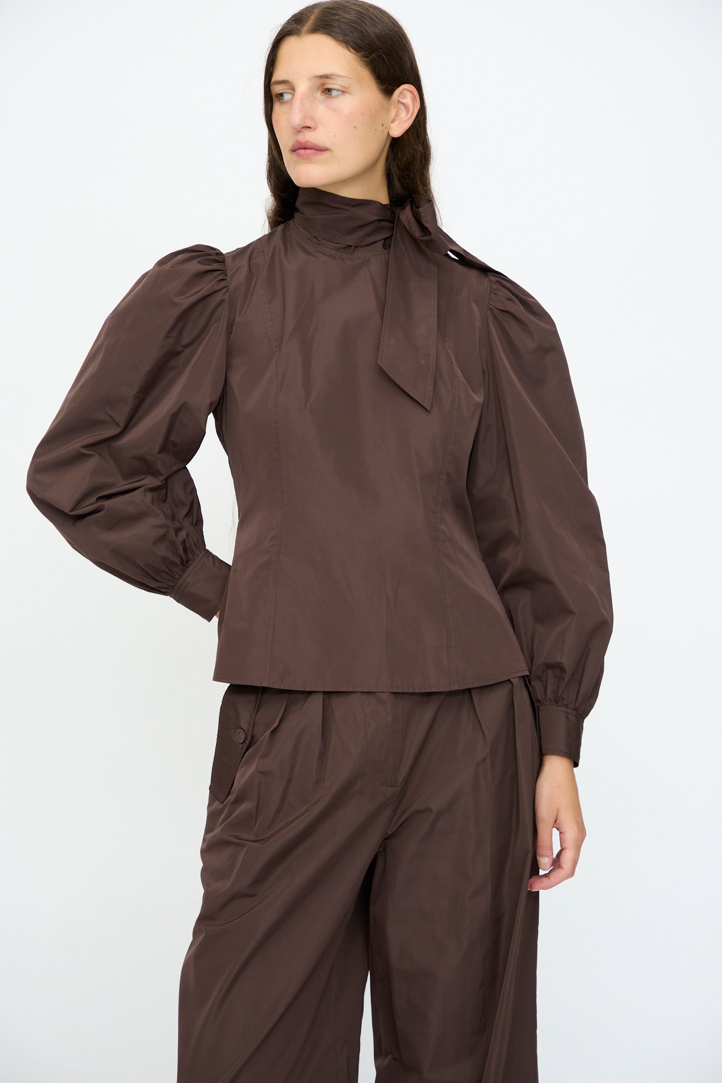 A person wearing the Ulla Johnson Giorgia Silk Blouse in Sequoia with matching brown pants, standing against a plain white background.