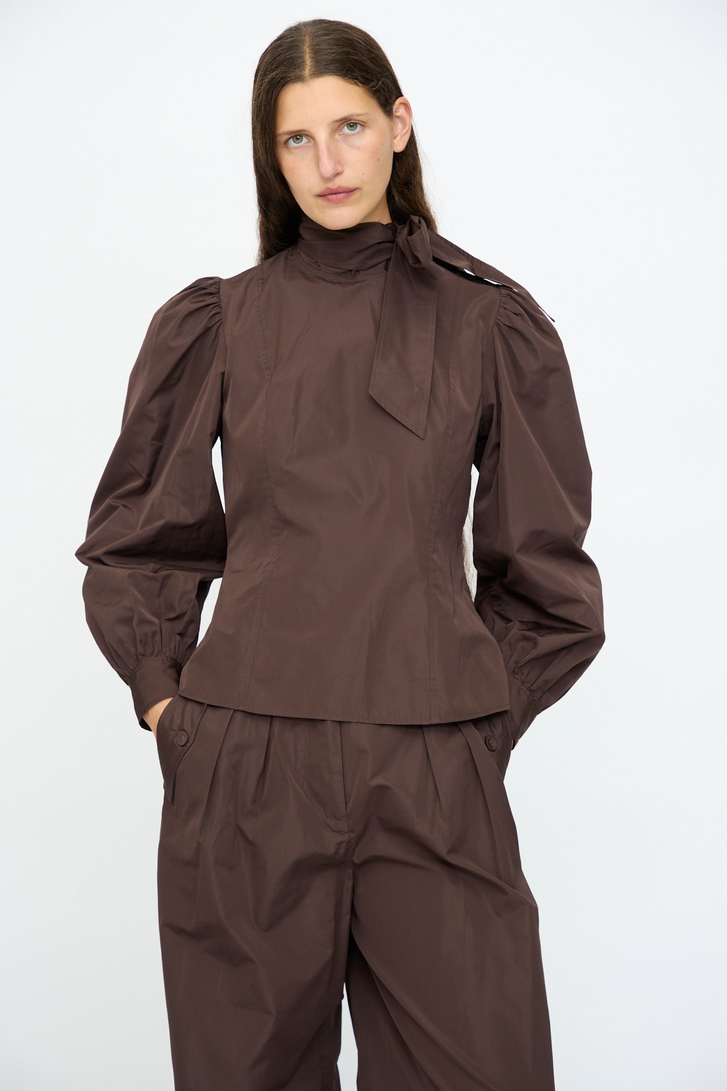 A woman wears the Ulla Johnson Giorgia Silk Blouse in Sequoia, featuring balloon sleeves and a bow neck, paired with matching high-waisted, wide-leg pants, standing against a plain white background.