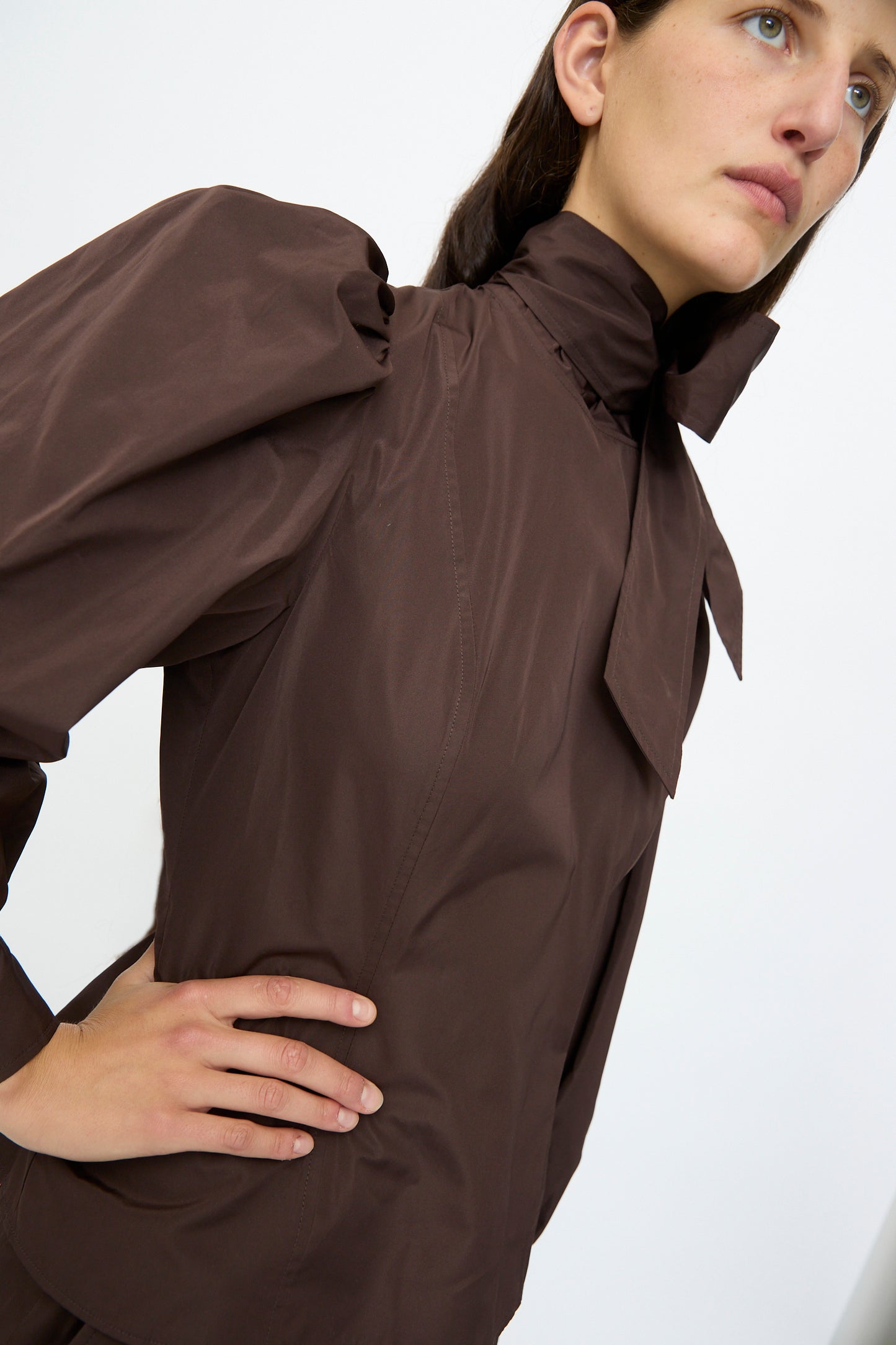 A person stands against a plain light background wearing the Ulla Johnson Giorgia Silk Blouse in Sequoia, featuring a high neck, bow detail at the collar, and balloon sleeves.